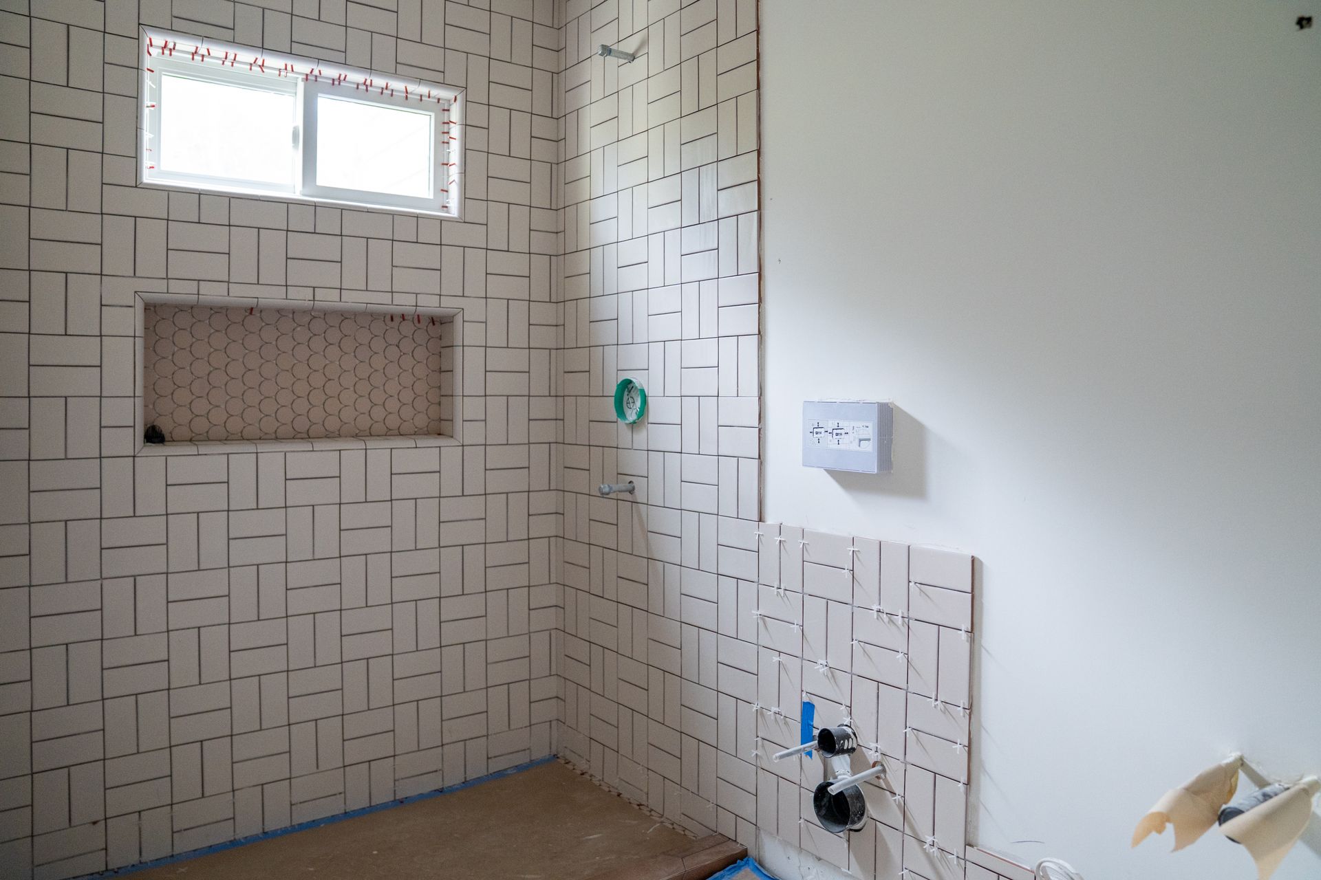 Empty tiled bathroom shower with small window, recessed shelf, and plumbing fixtures under construction