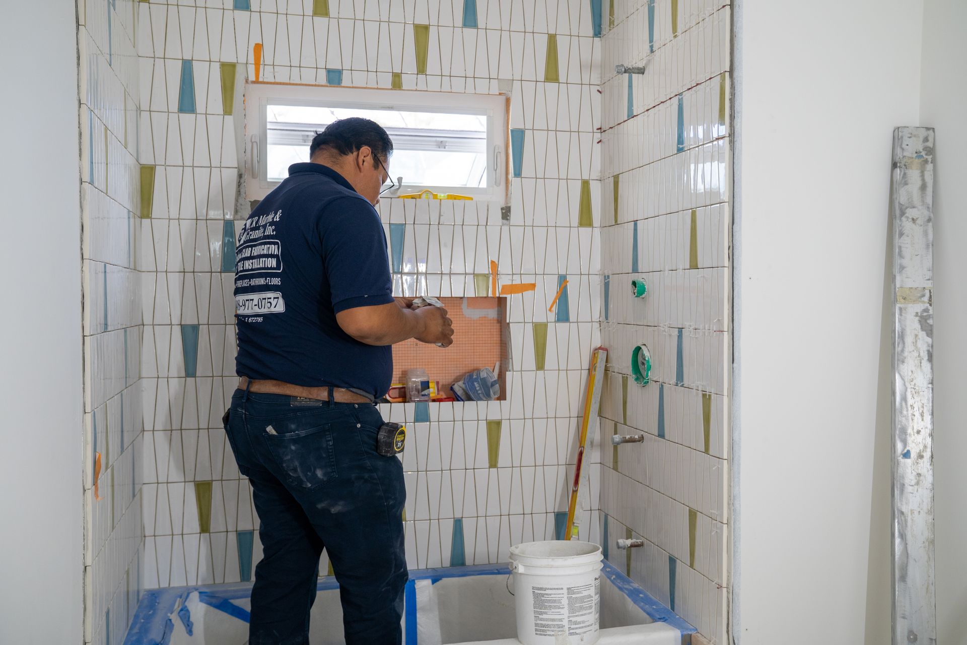 Worker tiling a bathroom wall beside a window, holding a tile and trowel.