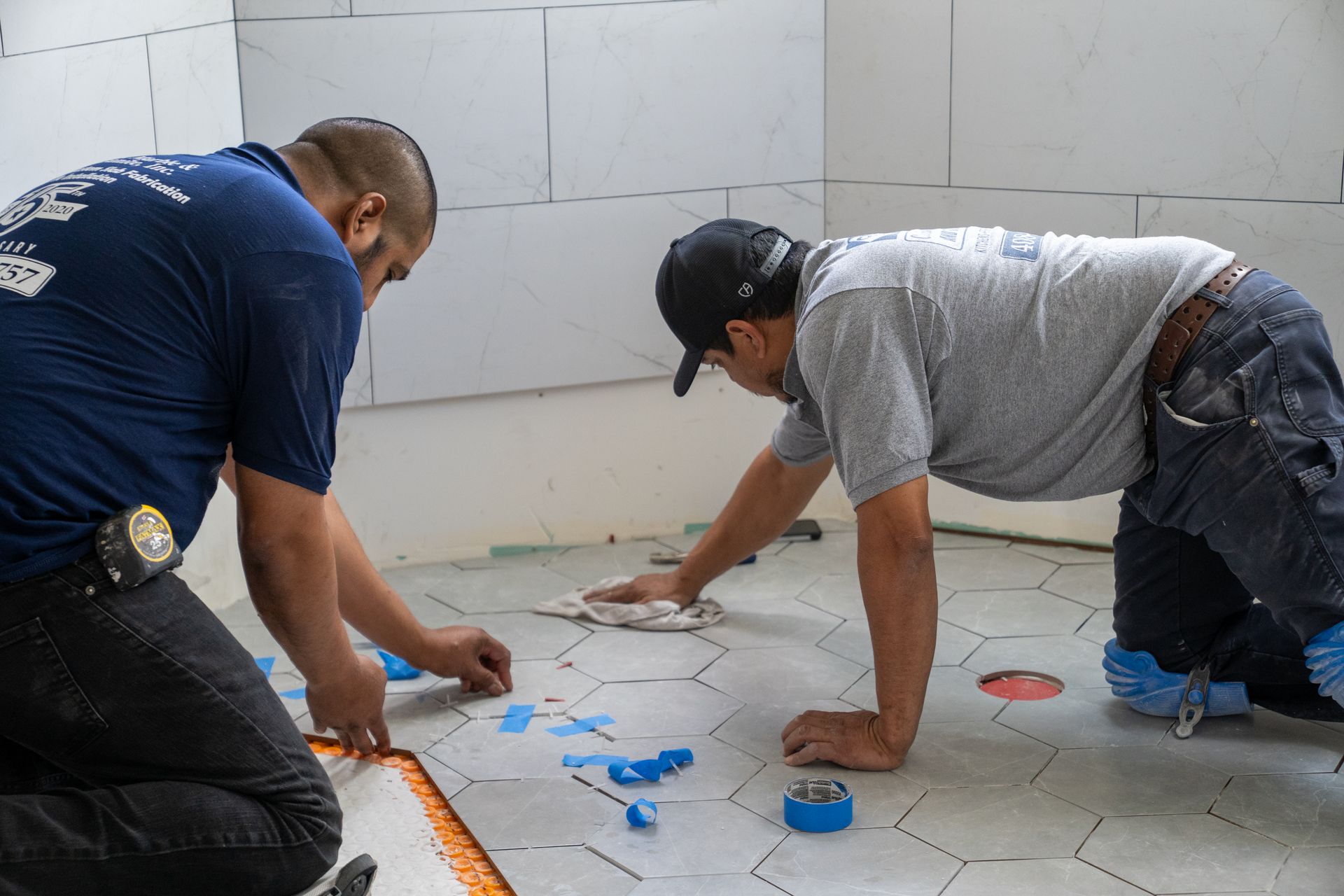 Two workers kneel on a tiled floor, applying blue spacers and markings during tile installation.