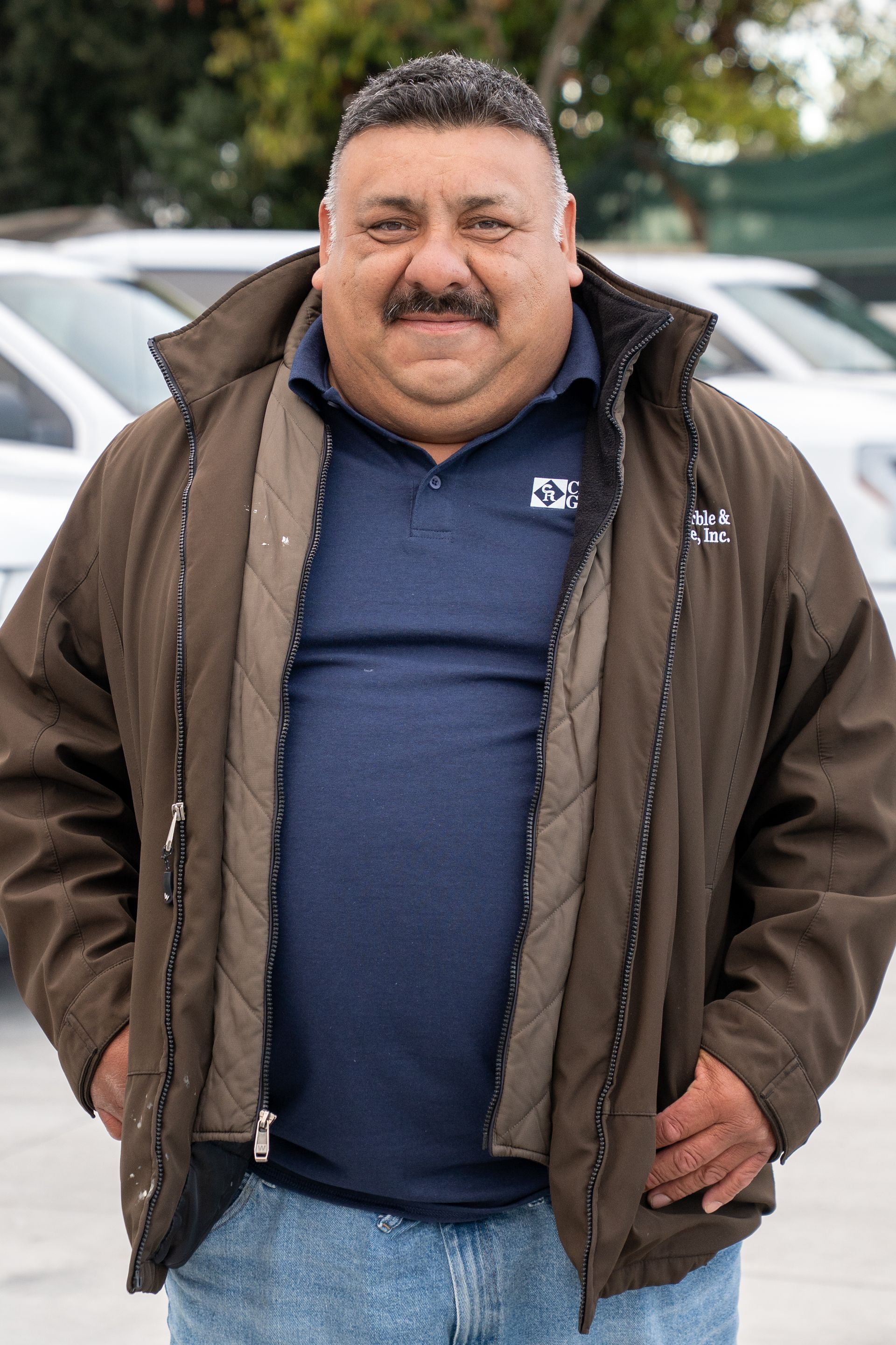 Man standing outdoors in a brown jacket and navy shirt, hands in pockets, with parked cars behind him