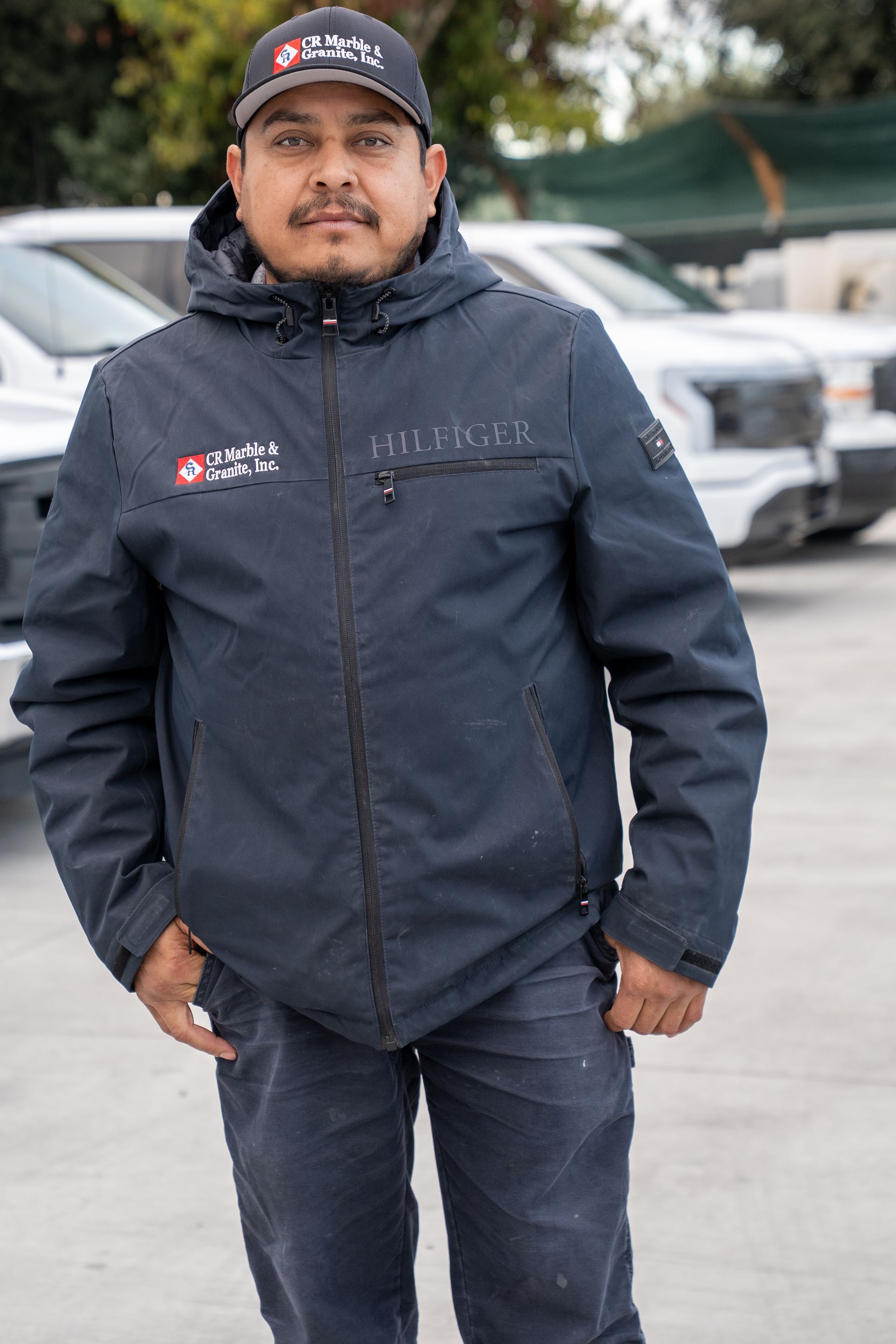 Man in a navy jacket and cap standing outdoors in a parking lot with vehicles behind him
