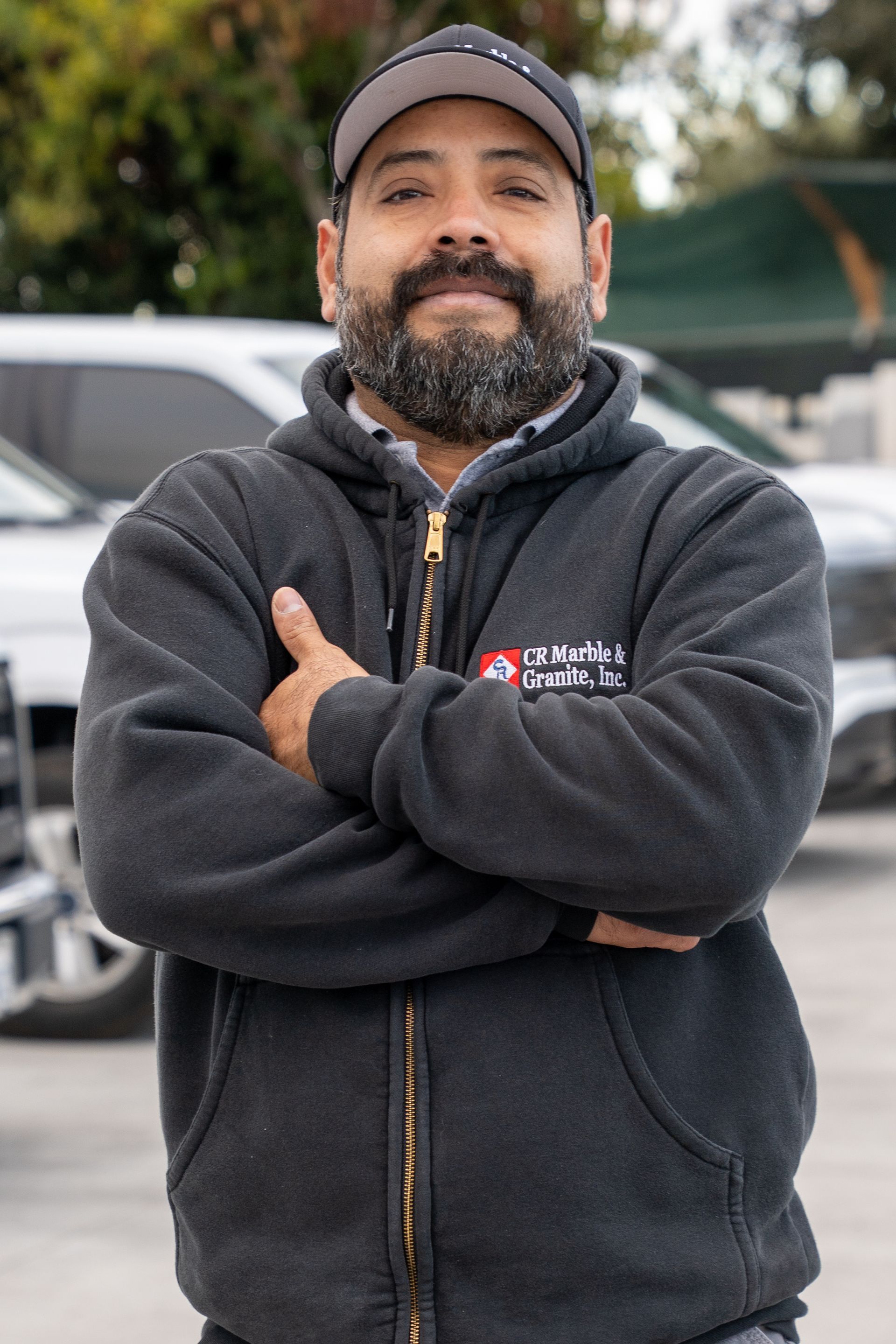 Man with folded arms wearing a dark hoodie and cap, standing outdoors in front of a parked vehicle.