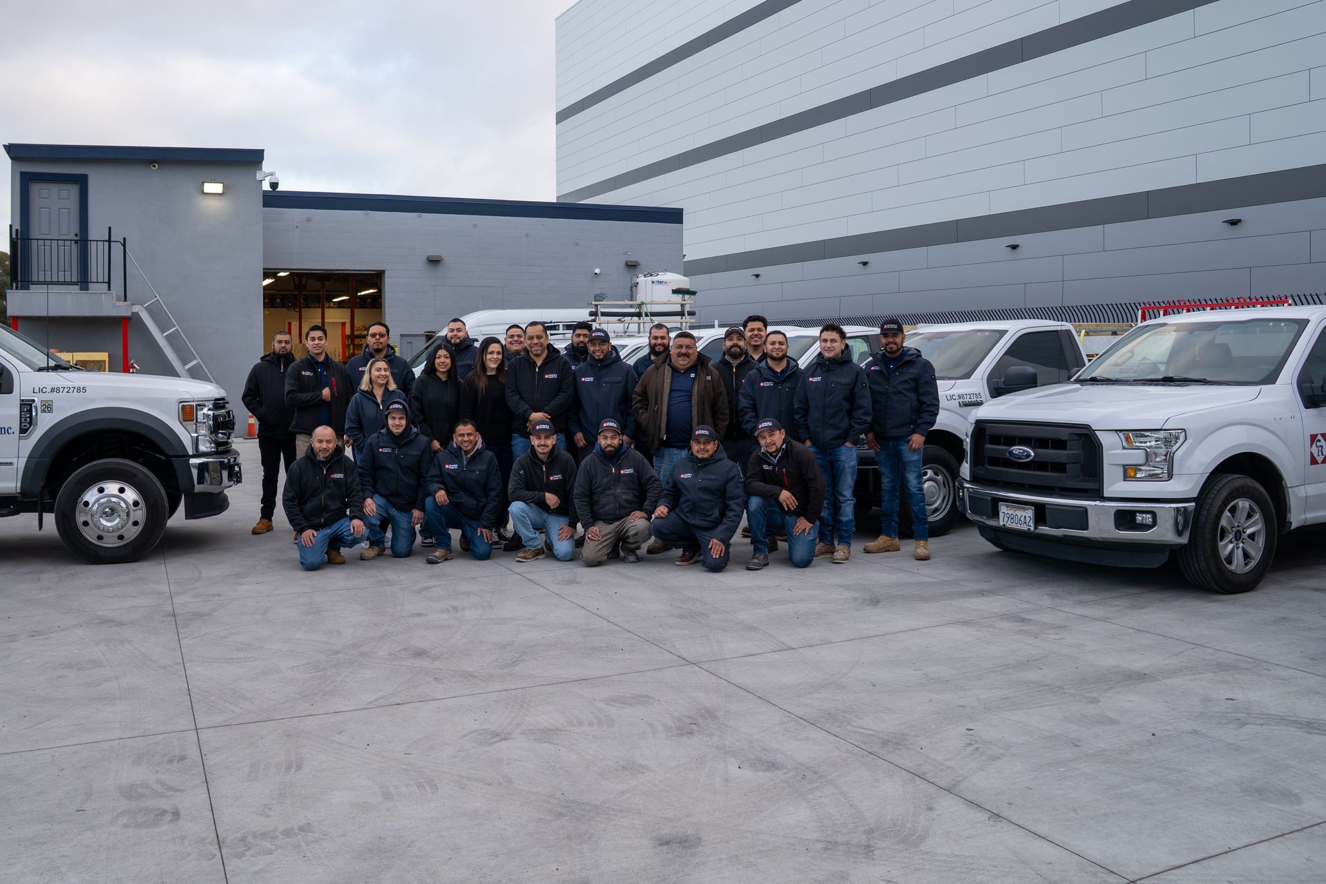 Group of uniformed workers posing outside a building beside two white trucks