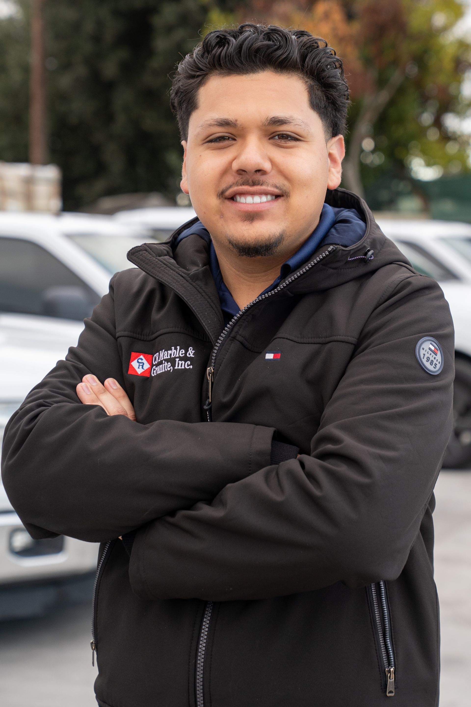 Smiling person in a black jacket standing with arms crossed outdoors beside parked cars.