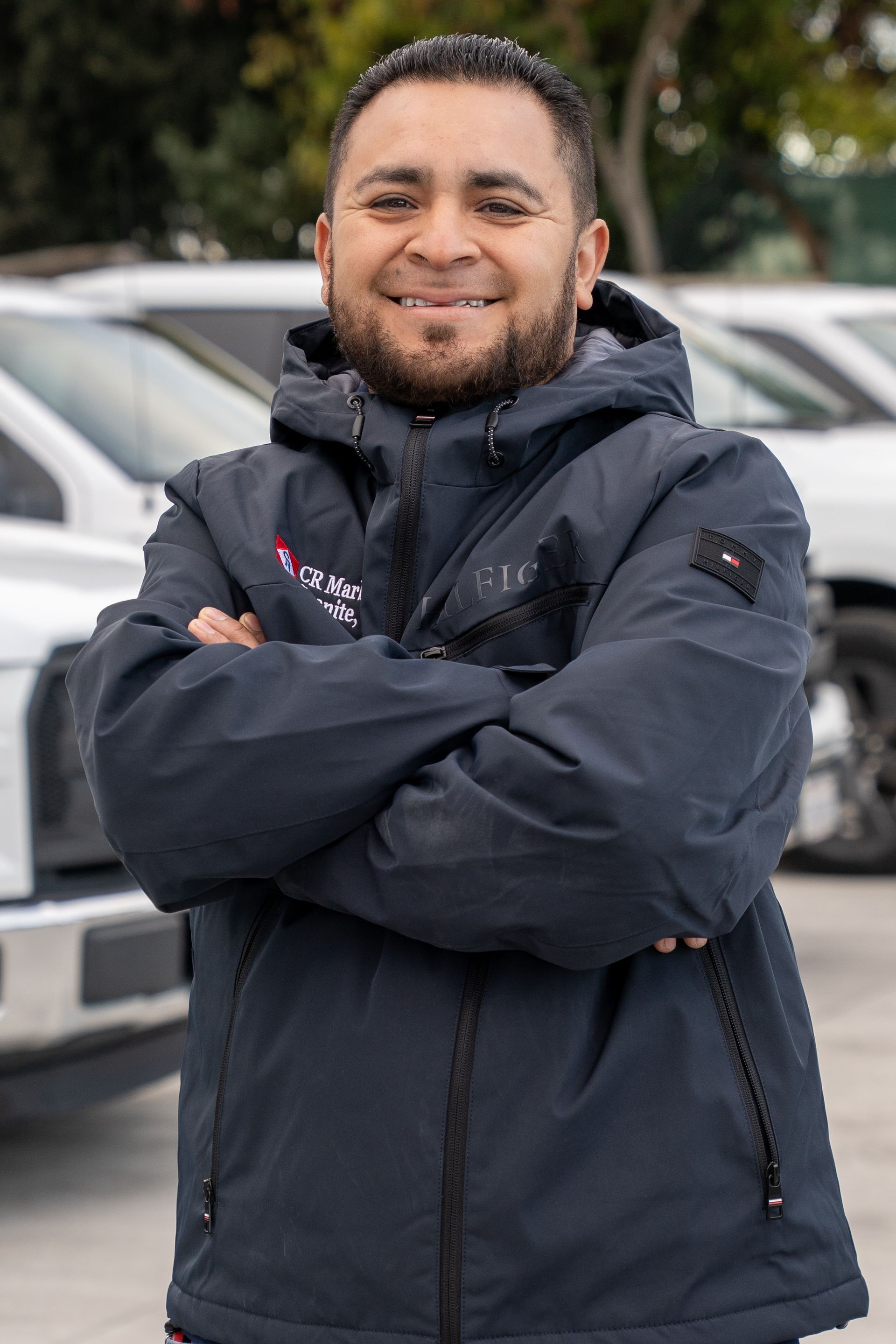 Smiling person in a dark jacket with arms crossed, standing outdoors in a parking lot.