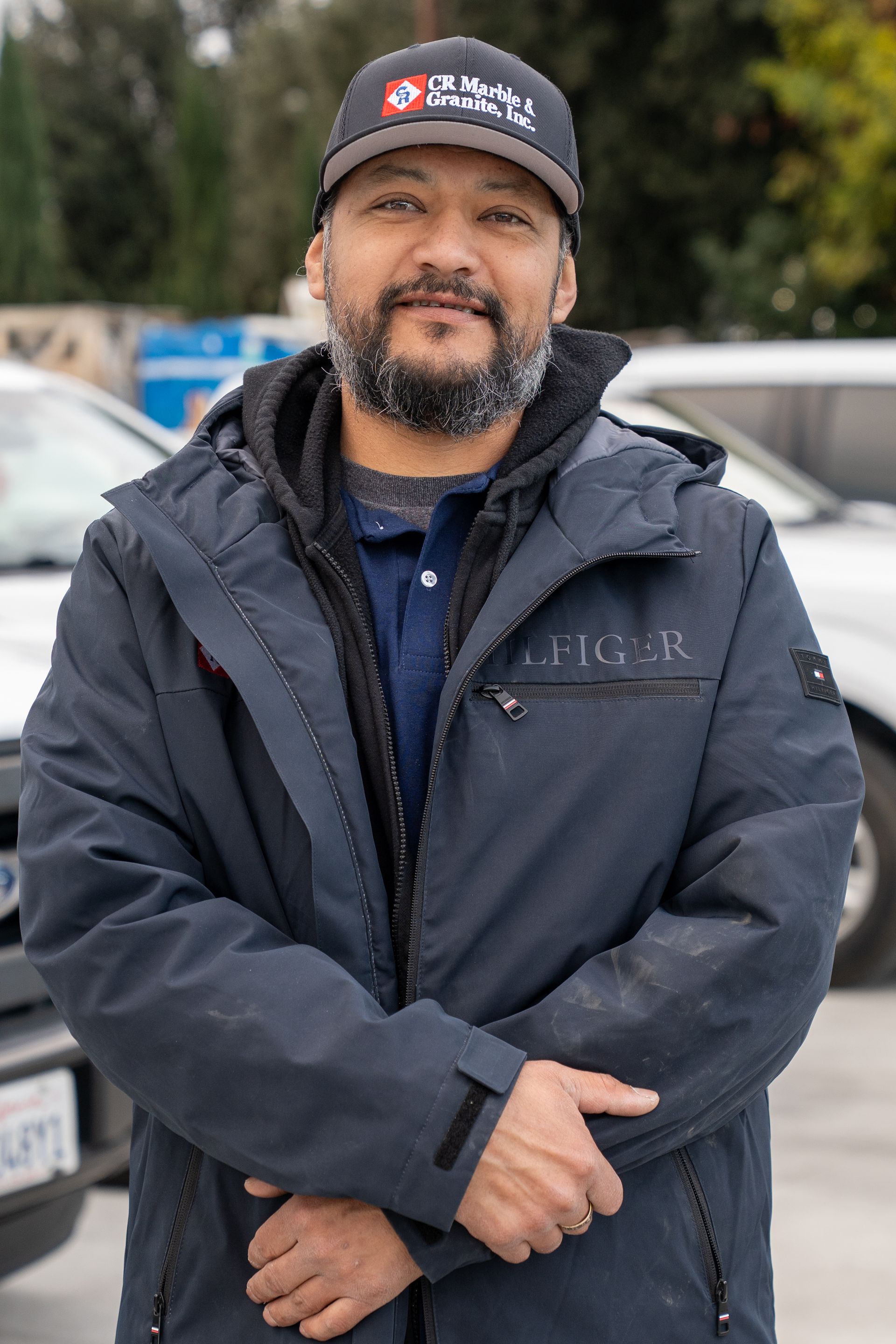 Man in a dark jacket and cap standing outdoors with arms crossed, in front of parked vehicles.