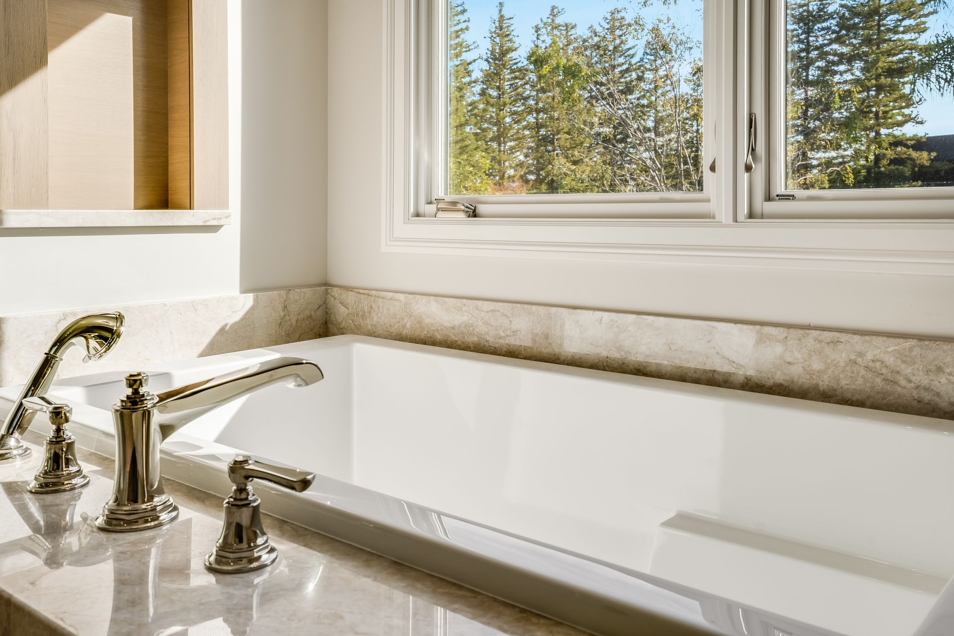 Bright white bathroom with a built-in bathtub, chrome faucet, and window overlooking trees.