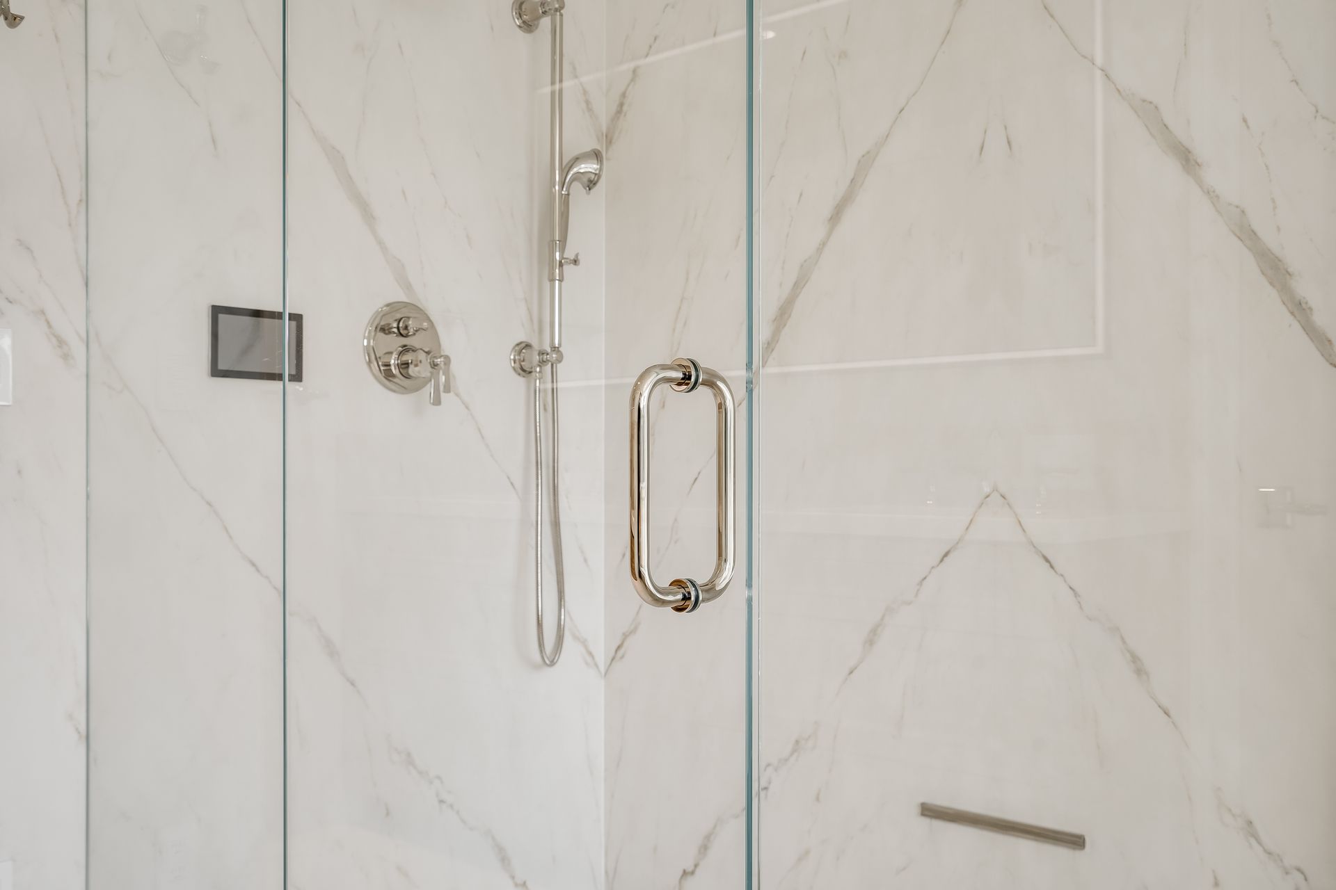 Glass shower door with chrome handle and shower fixtures in a white marble-tile bathroom