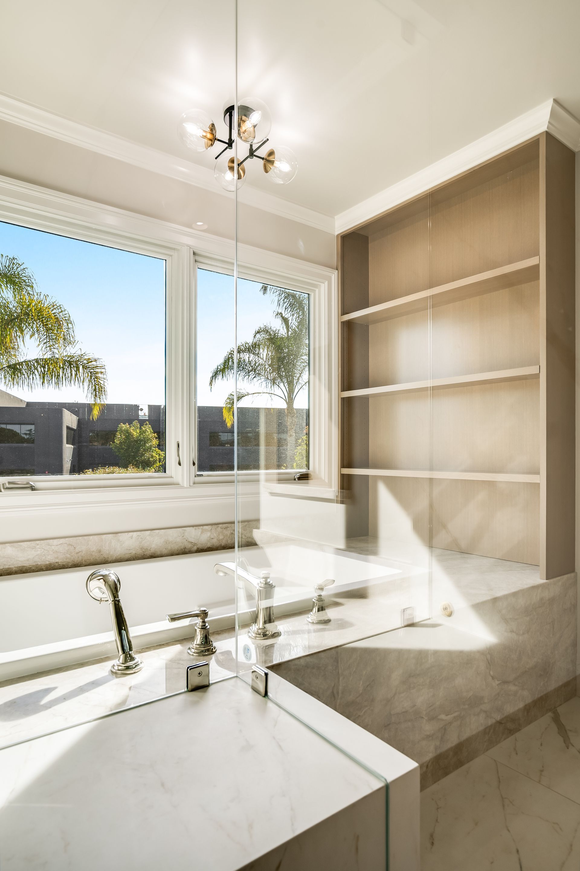 Bright modern bathroom with a large window, white vanity, beige built-in shelves, and a chandelier.