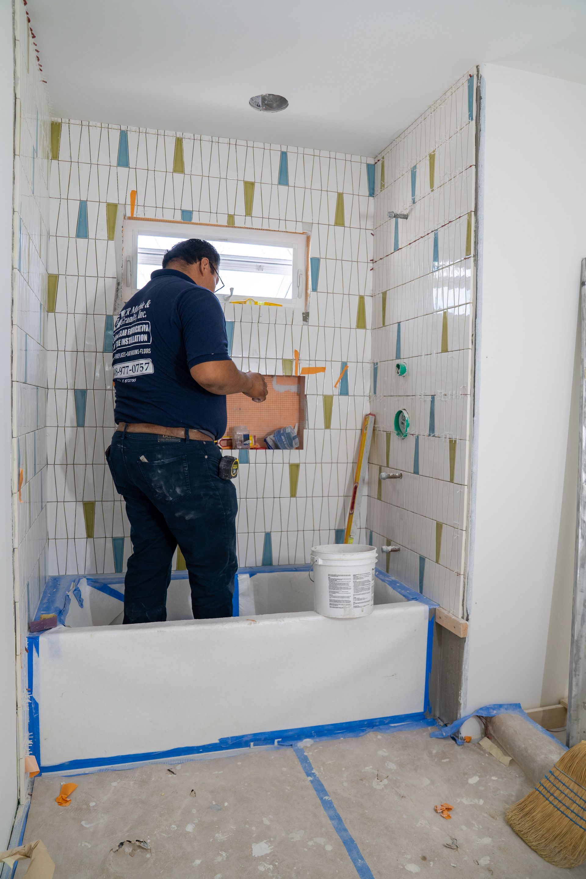 Man tiling a bathroom, standing in the bathtub. Walls are patterned, with a small window and recessed shelf.