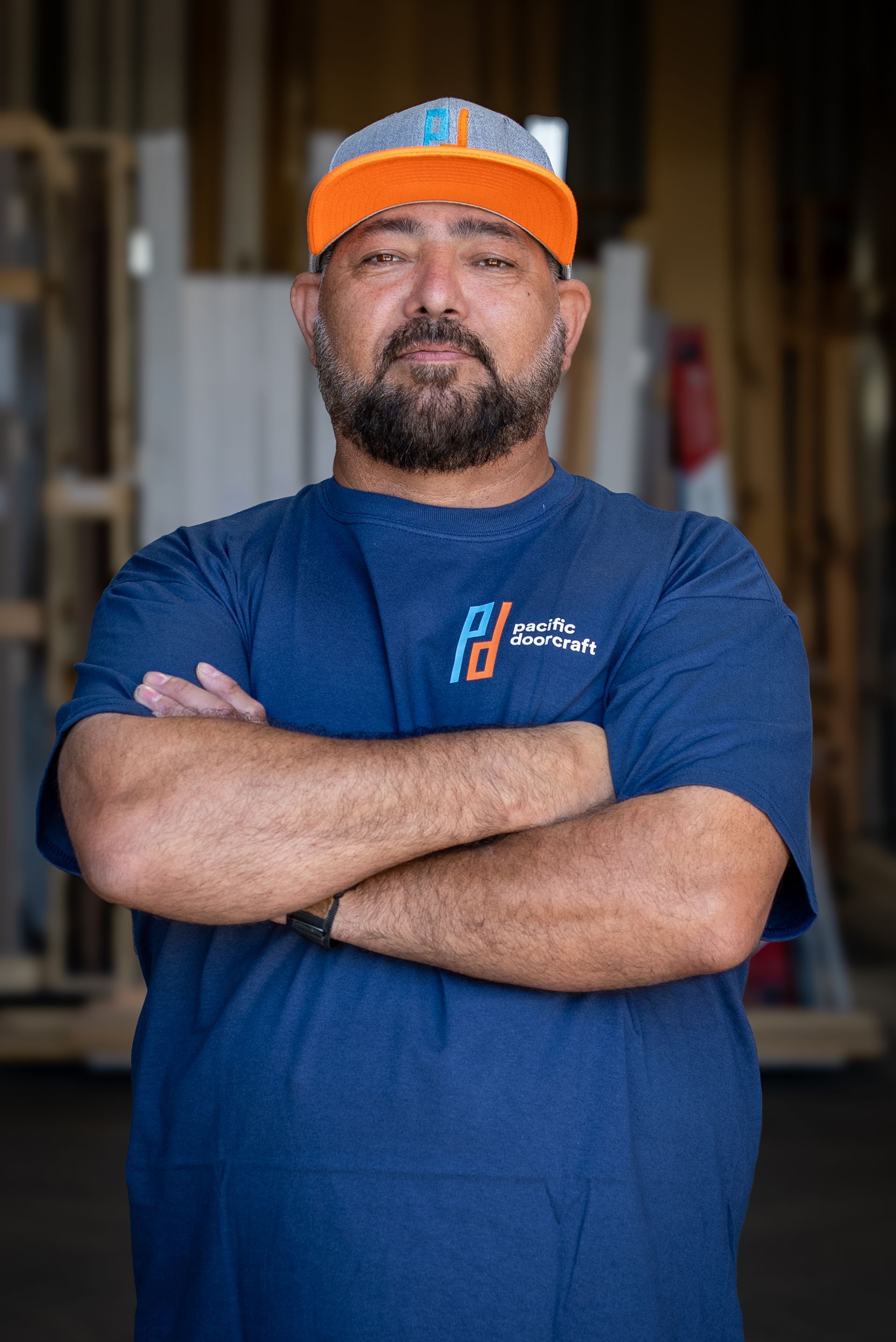 Person in an orange cap and blue T-shirt stands with arms crossed in a workshop.
