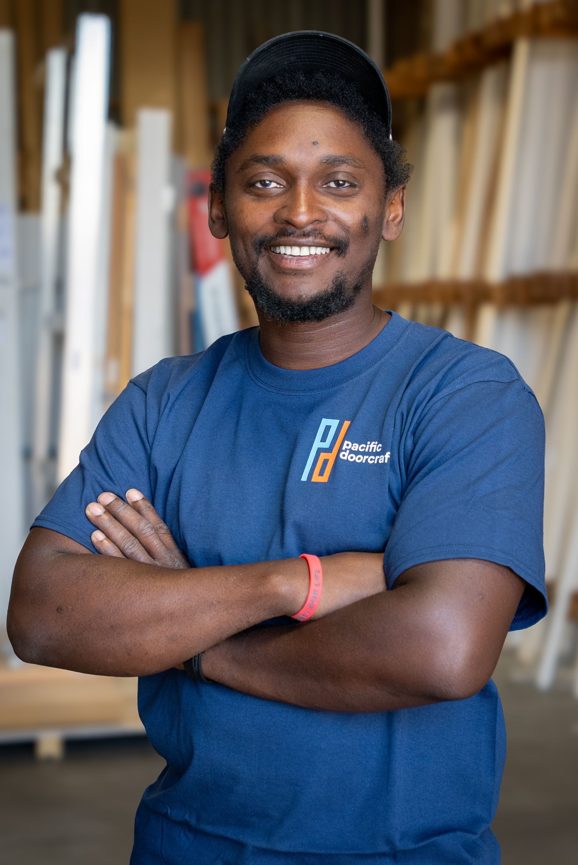 Smiling person in a blue T-shirt standing with arms crossed in a workshop or storage area