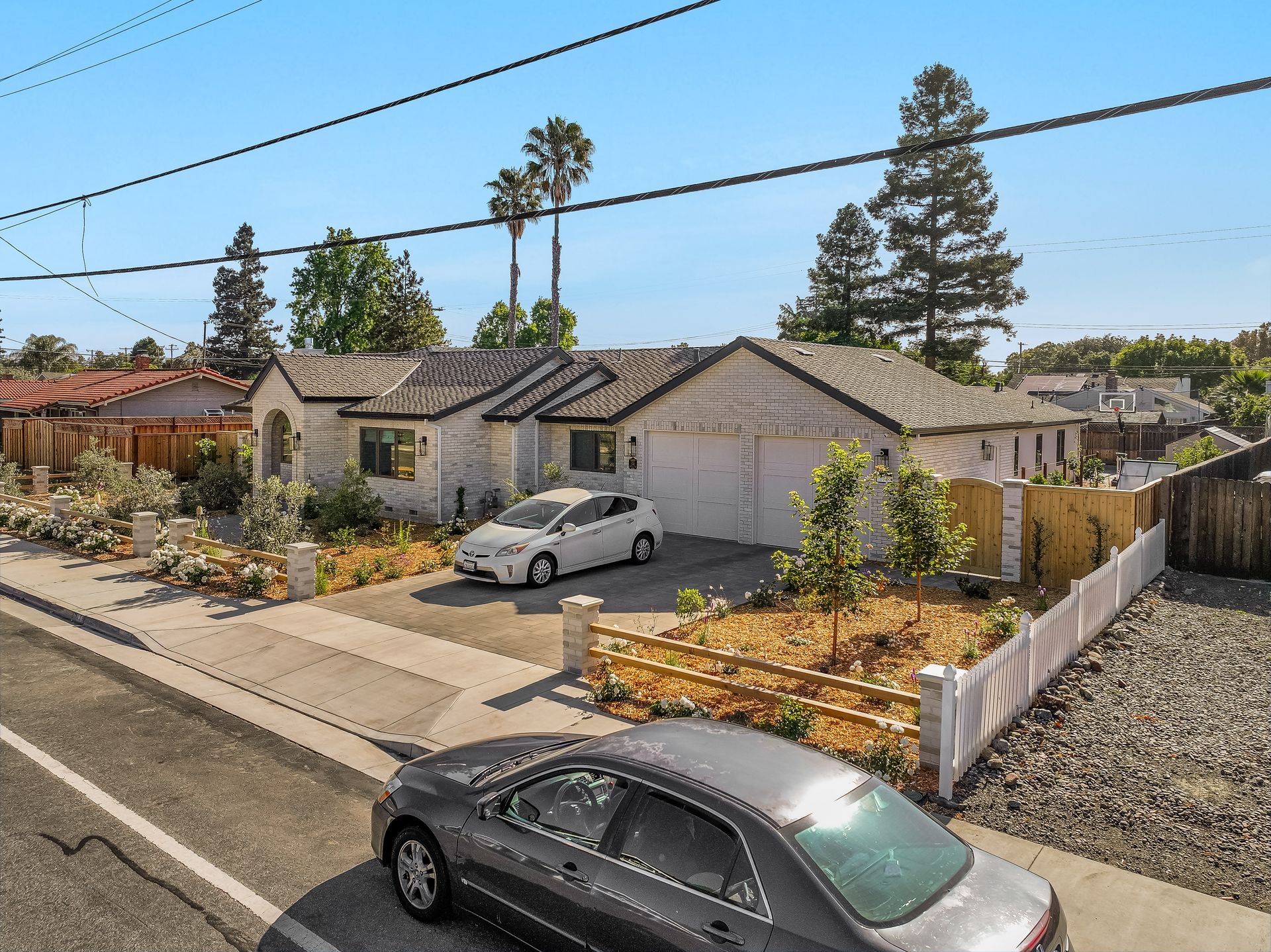 Street view of a small house with a damaged roof, parked cars, and a white fence under a clear blue sky