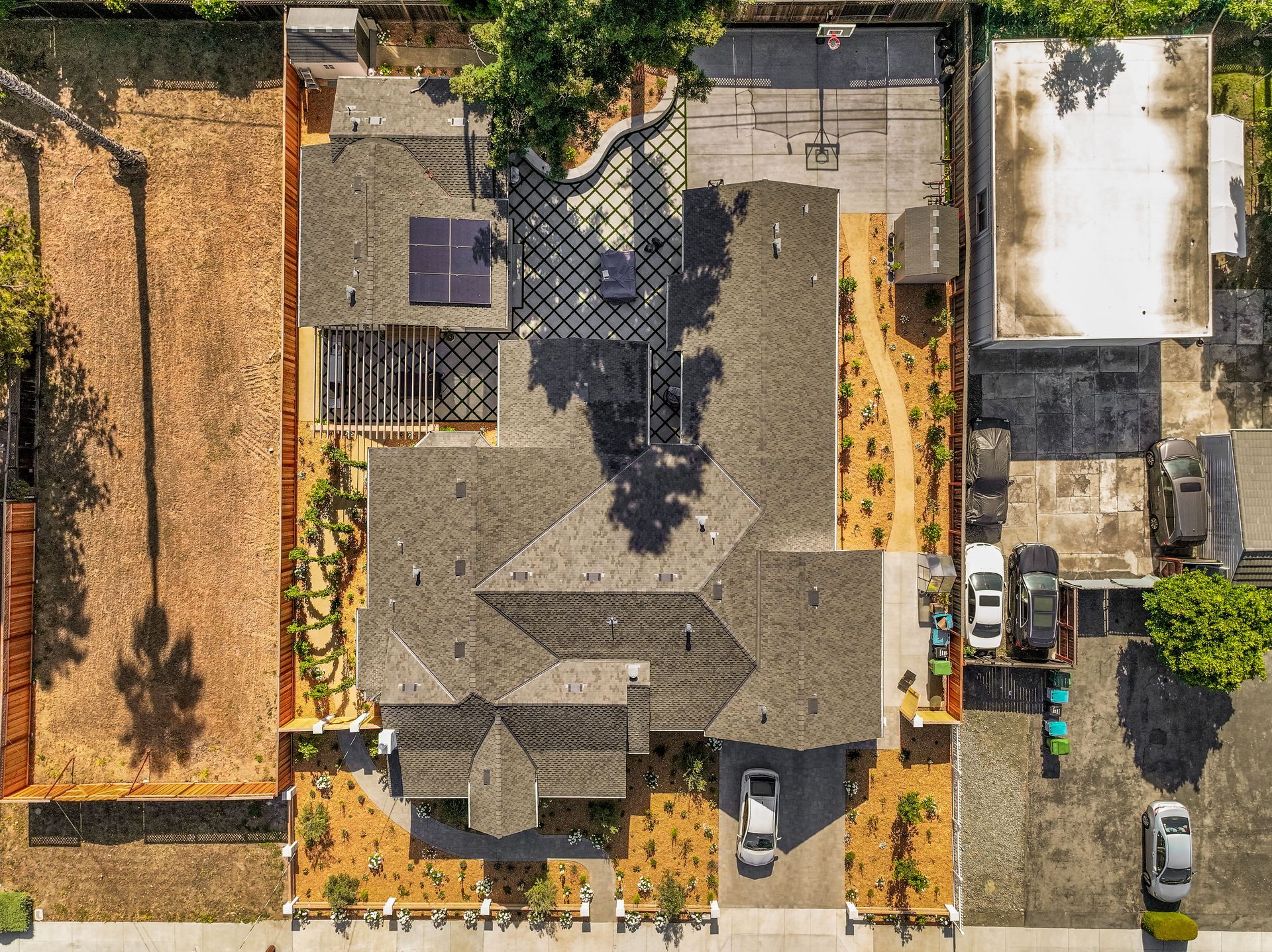 Aerial view of a house with a gray roof, driveway, parked car, and surrounding trees and neighboring lots.