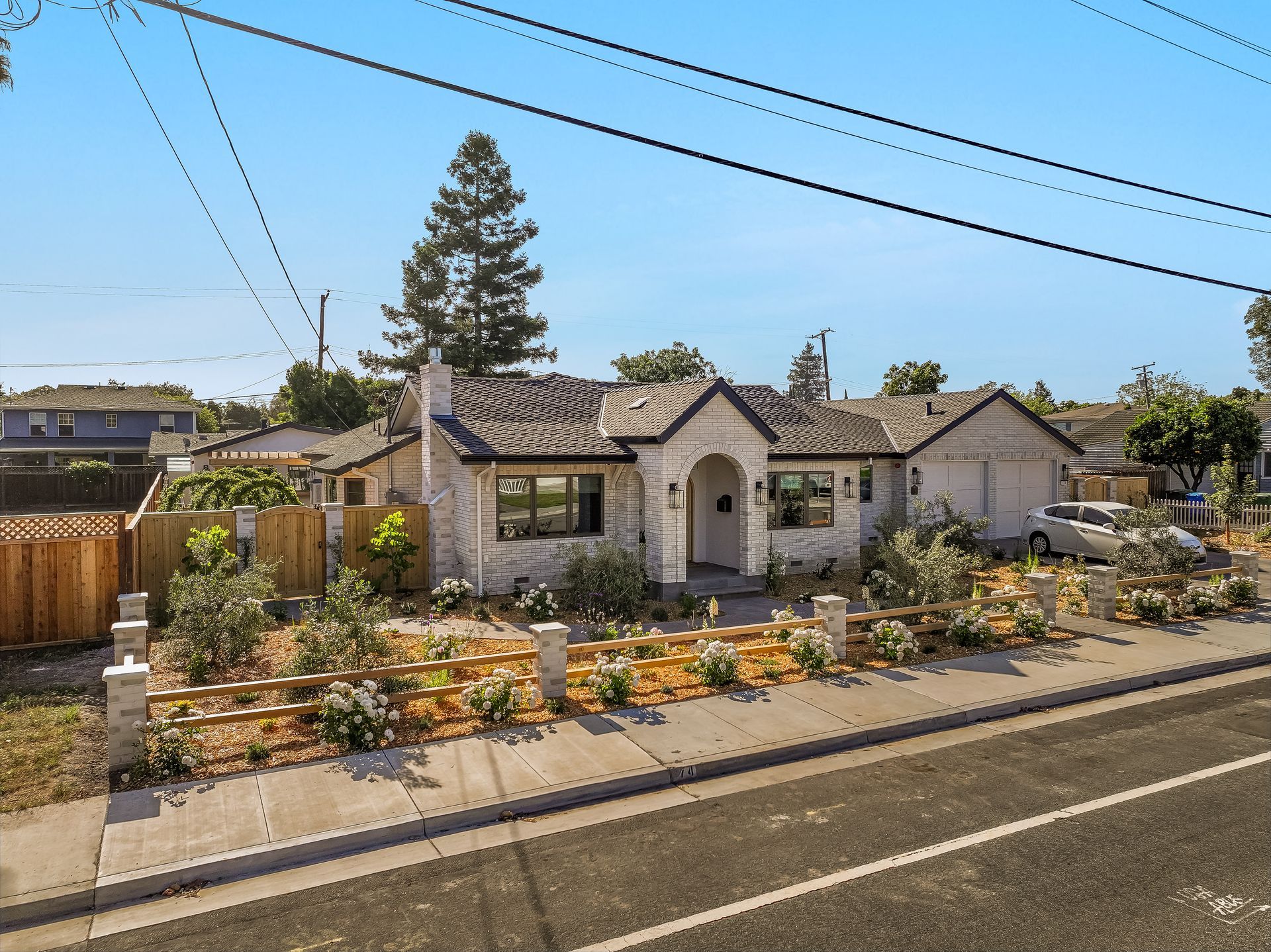 Single-story house with a stone facade, white front gate, and colorful flower garden along a suburban street.