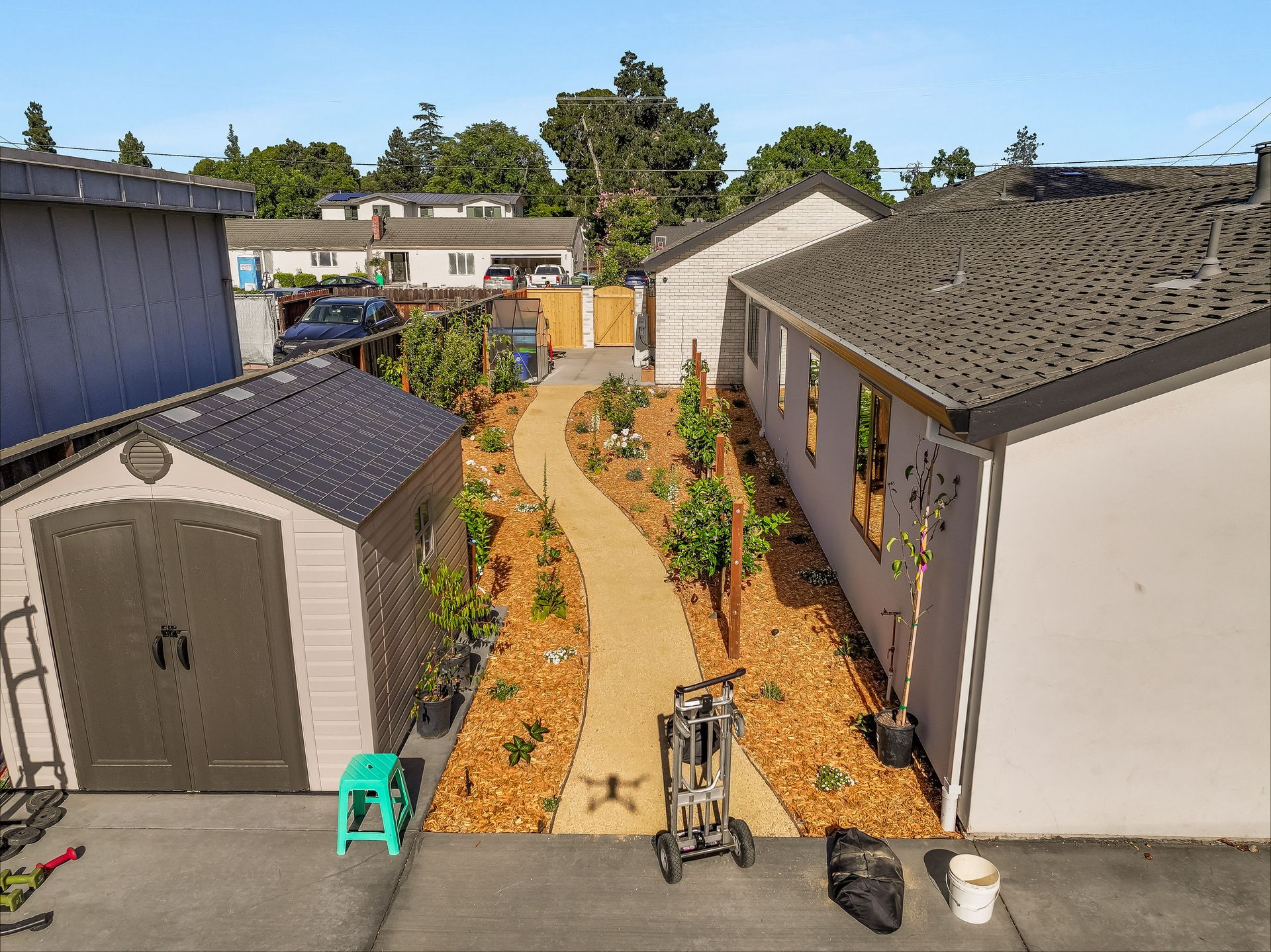 Aerial view of a narrow garden path between two sheds, lined with mulch and plants.