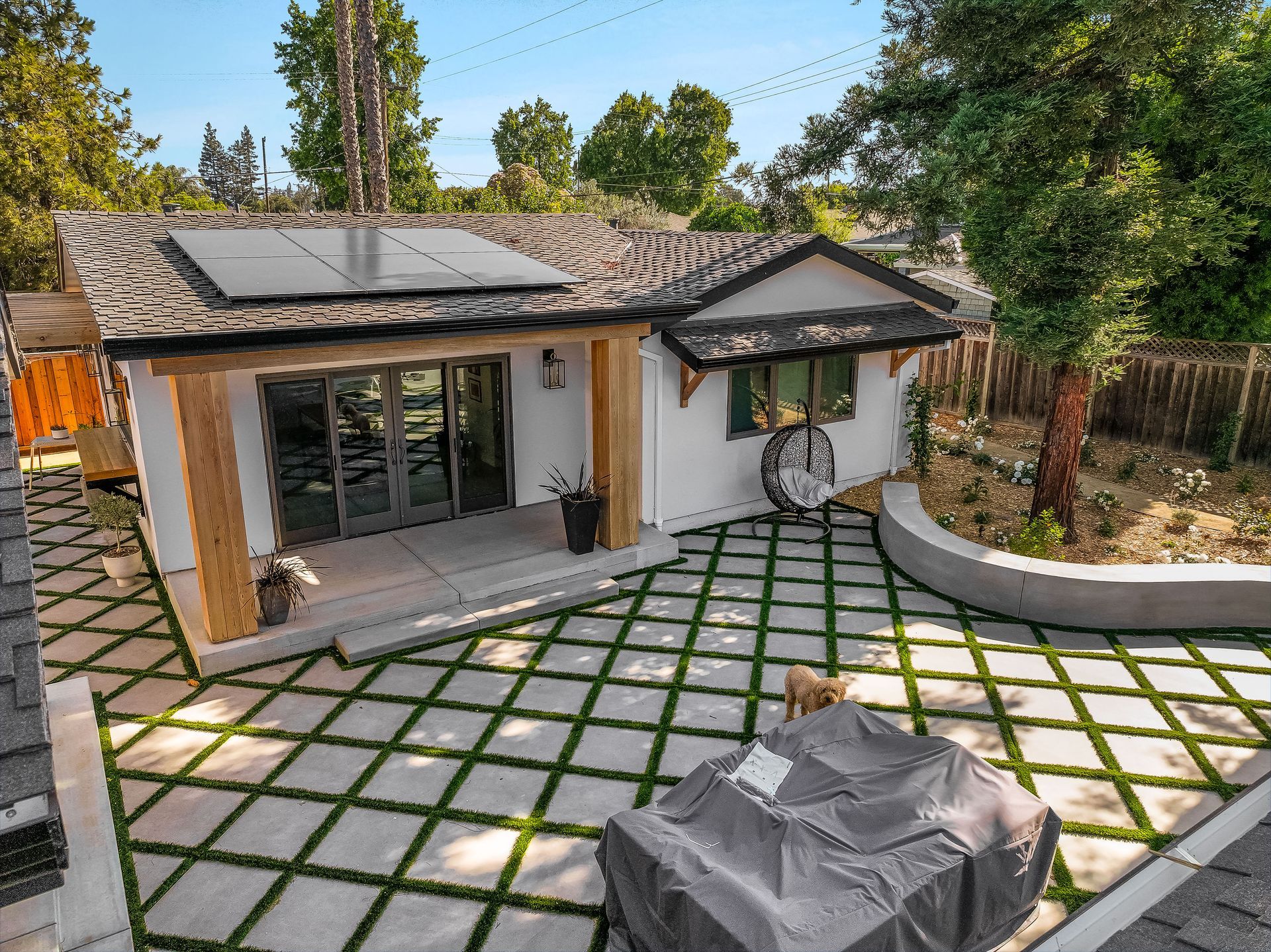 Backyard patio with modern house, tiled courtyard, covered grill, and trees.