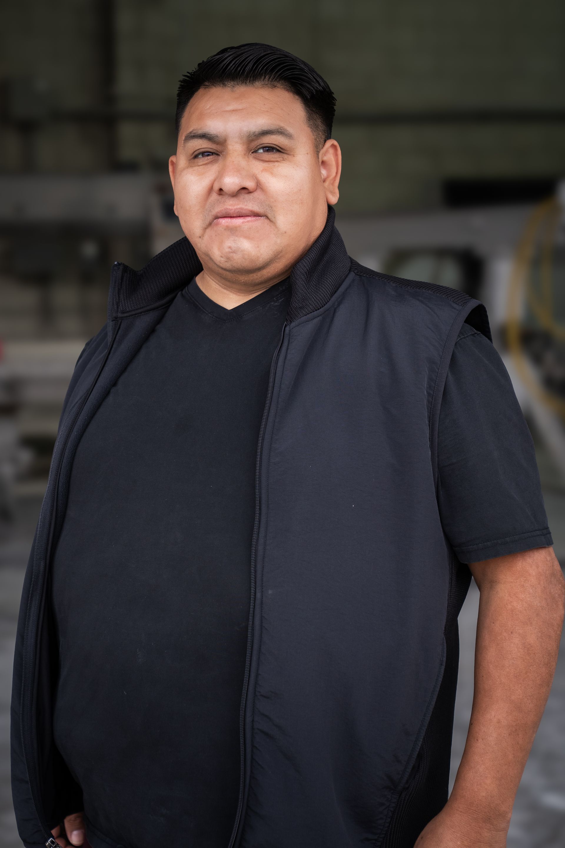 Person in a black shirt and vest standing indoors against a blurred background.