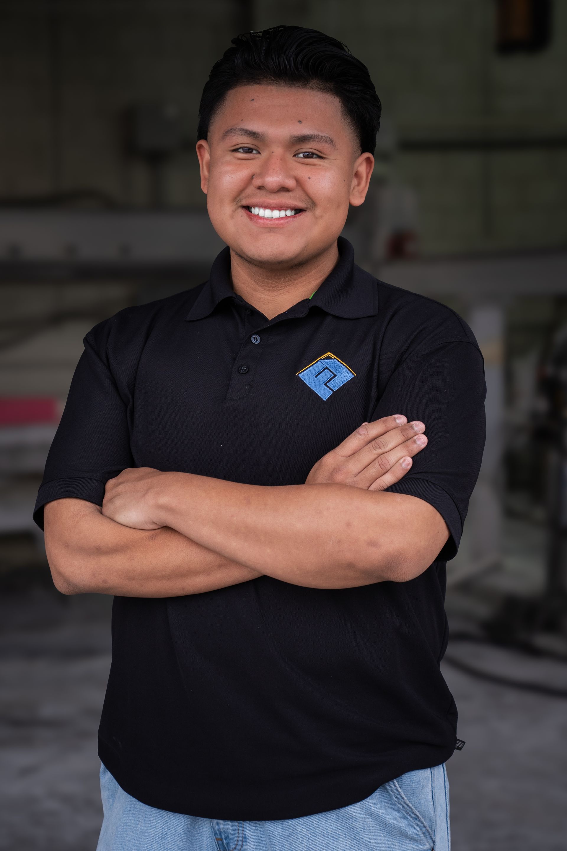 Smiling person in black polo shirt with arms crossed, standing indoors against a blurred background