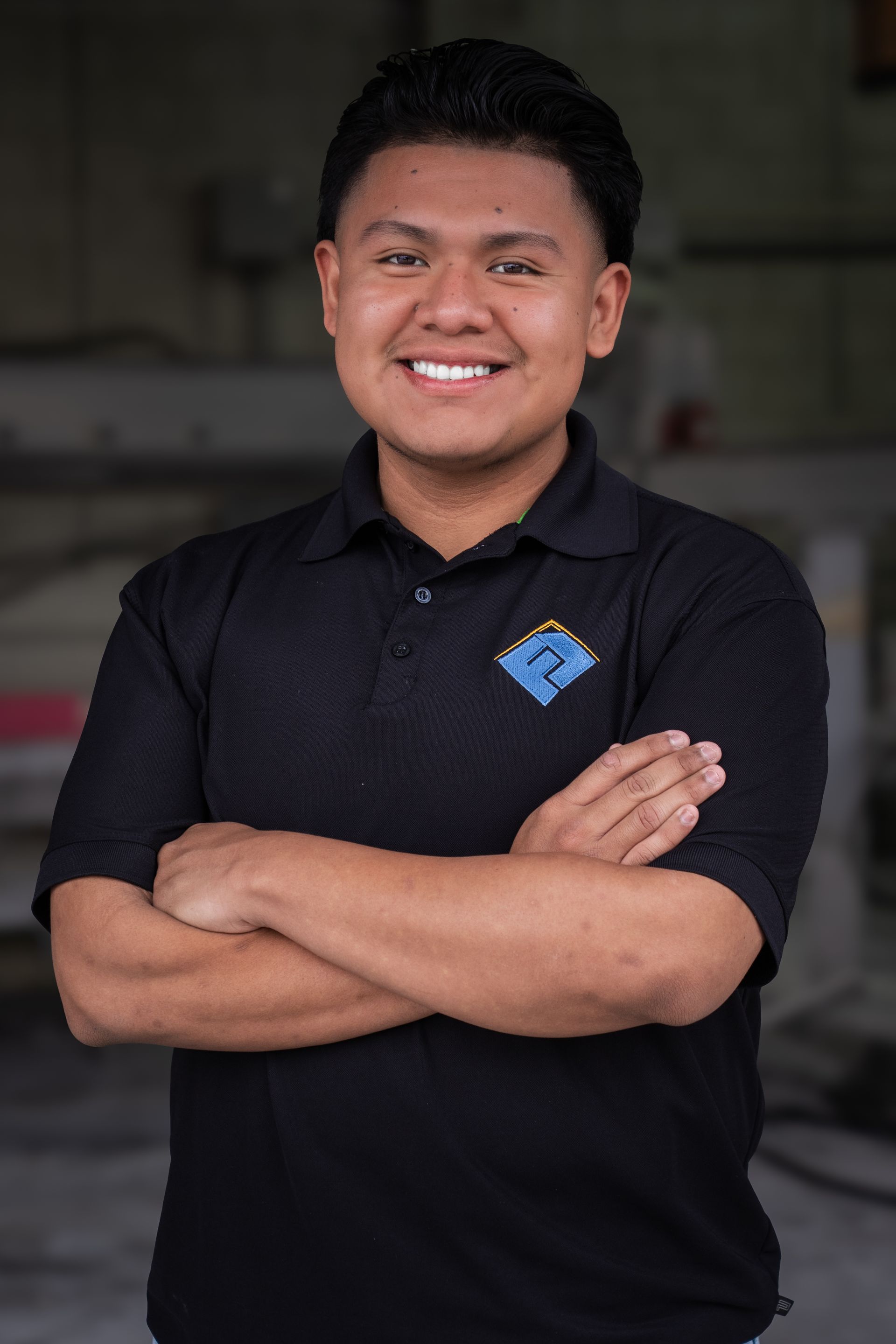 Smiling person in a black polo shirt with arms crossed, standing indoors against a blurred background