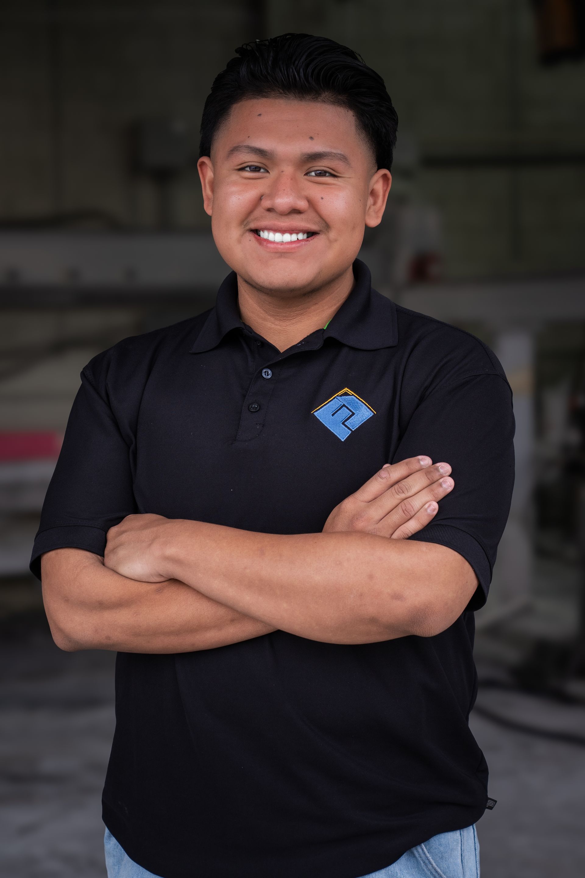 Smiling person in a black polo shirt with arms crossed, standing in a dim indoor setting.