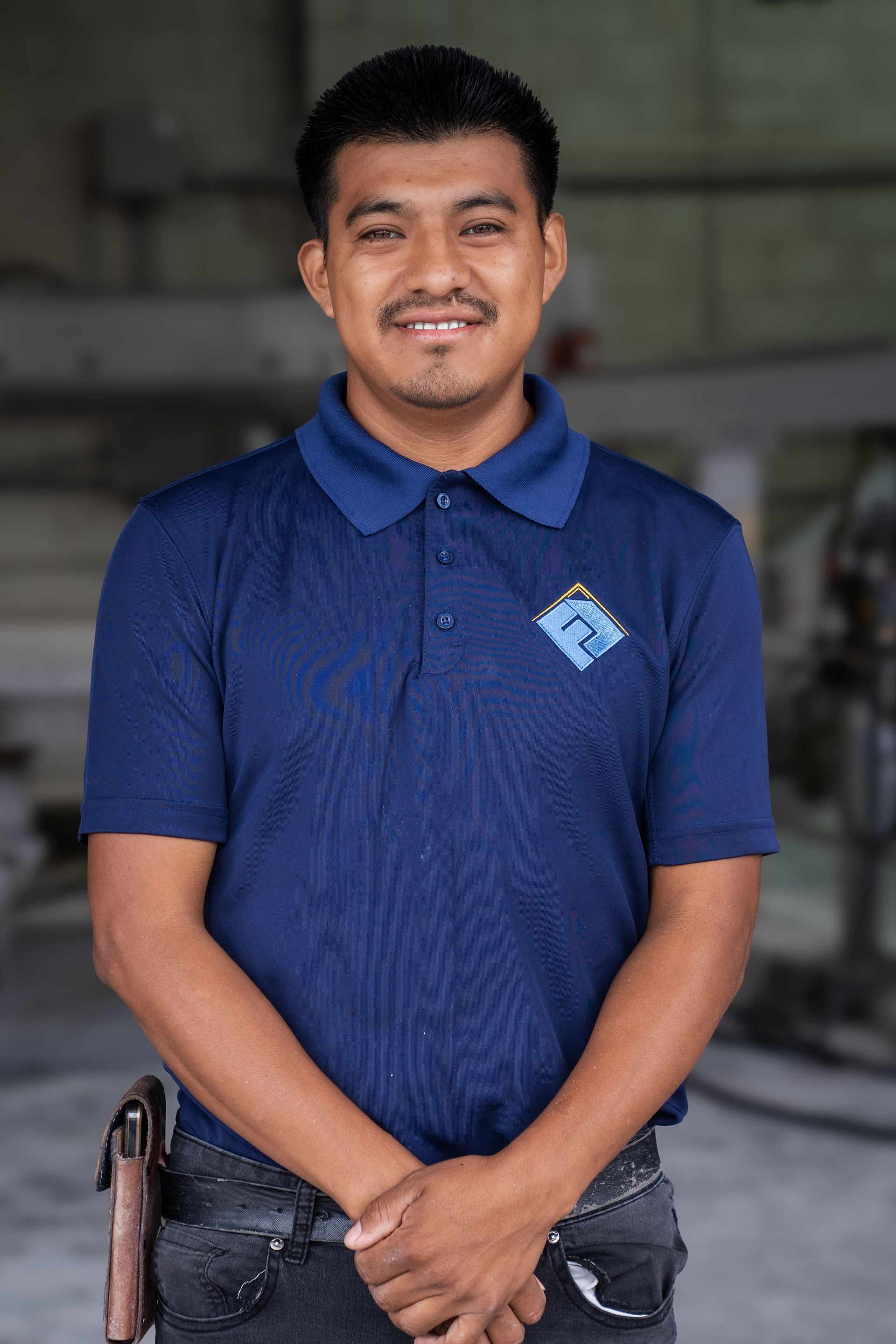 Smiling person in a blue polo shirt standing indoors with hands clasped in front of them