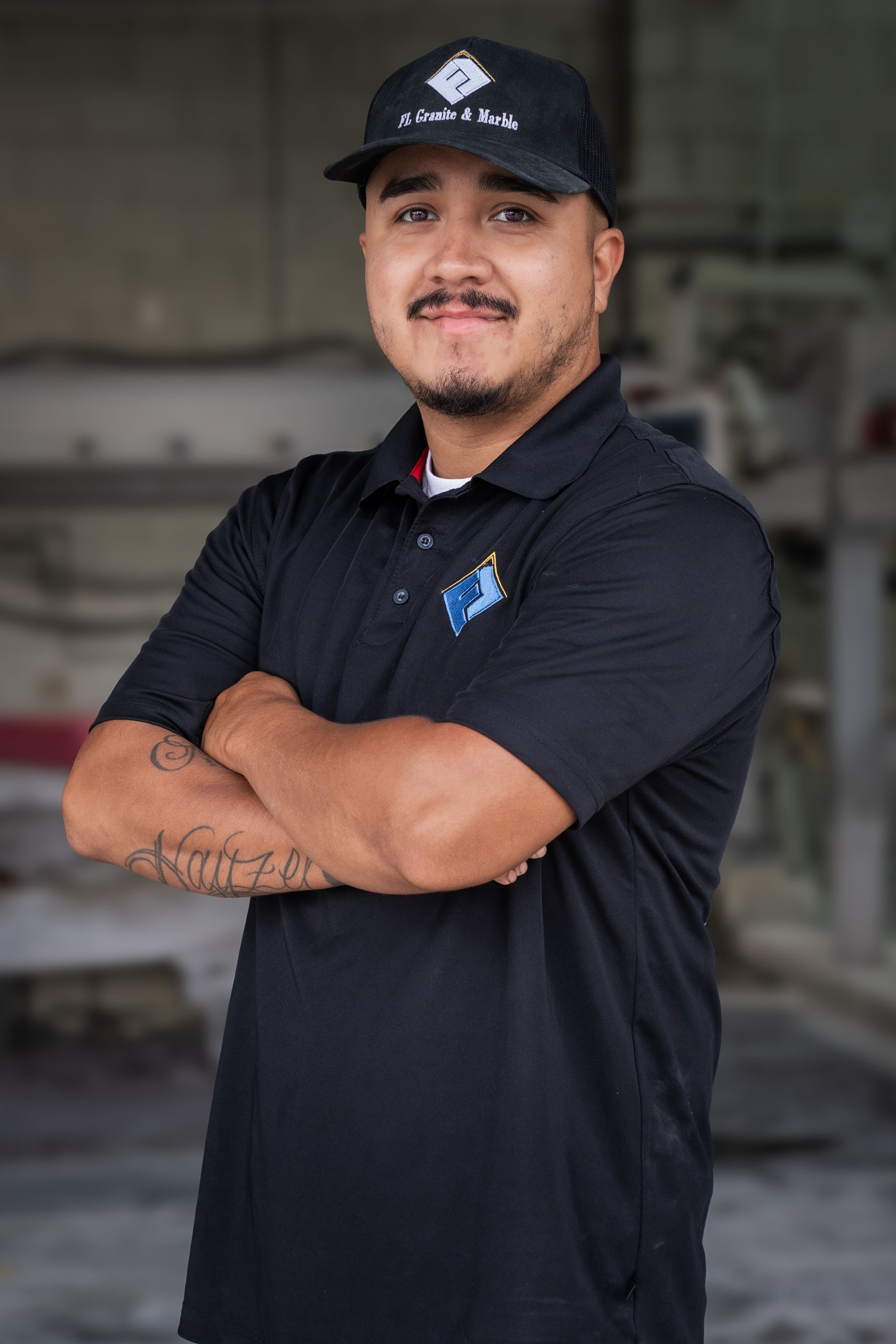 Man in black work uniform and cap, arms crossed, standing in an industrial setting.