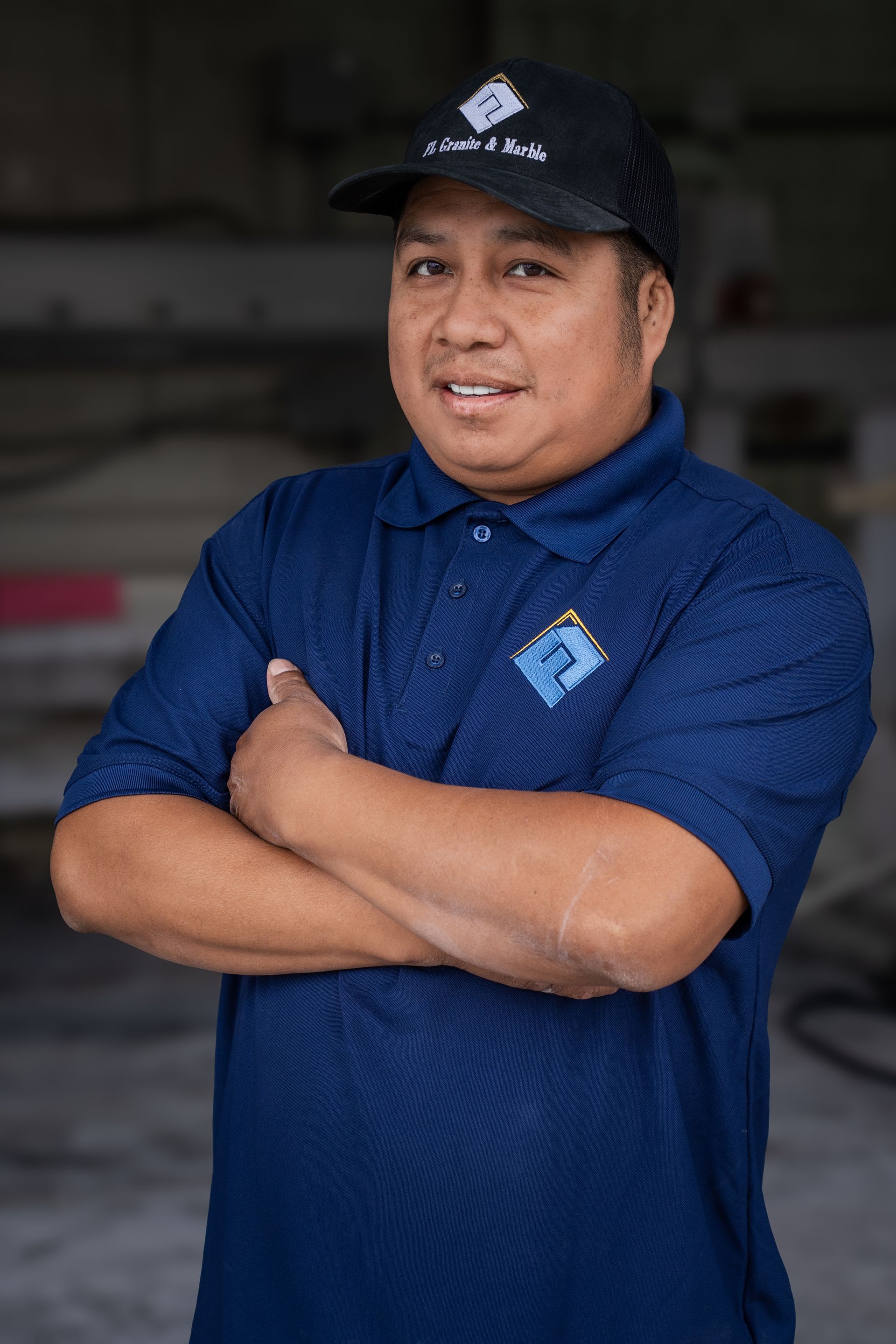 Man in a navy polo and black cap stands with arms crossed in a workshop, smiling at the camera.