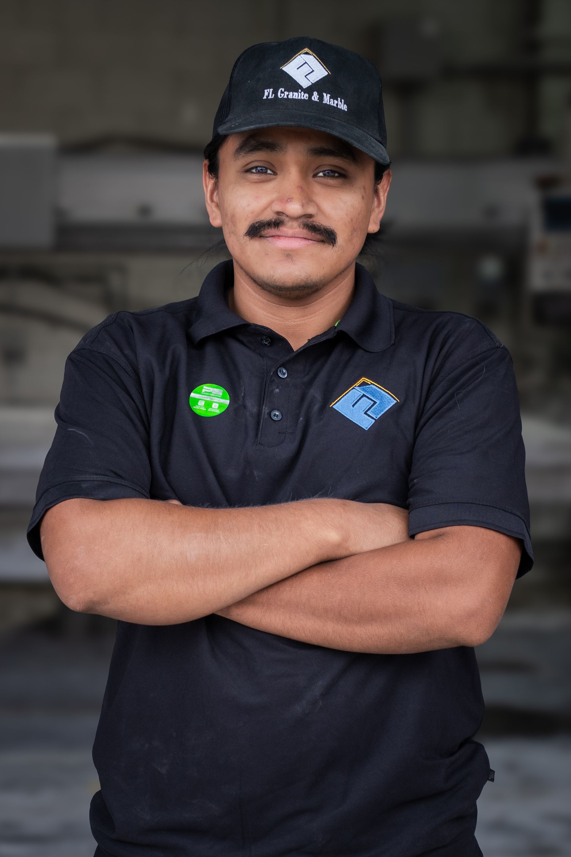 Man in black polo and cap stands with arms crossed in a garage, smiling slightly.