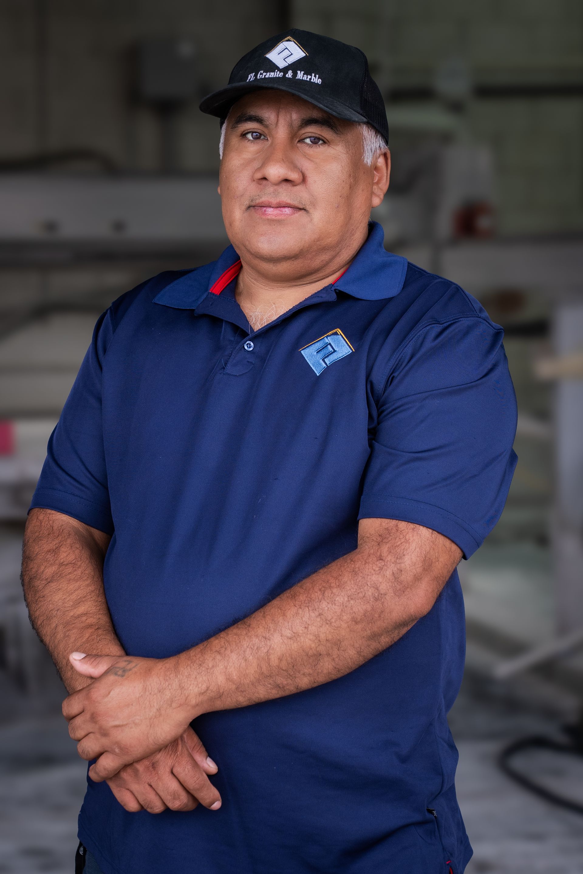 Man in a navy polo and black cap standing with arms crossed in an industrial setting