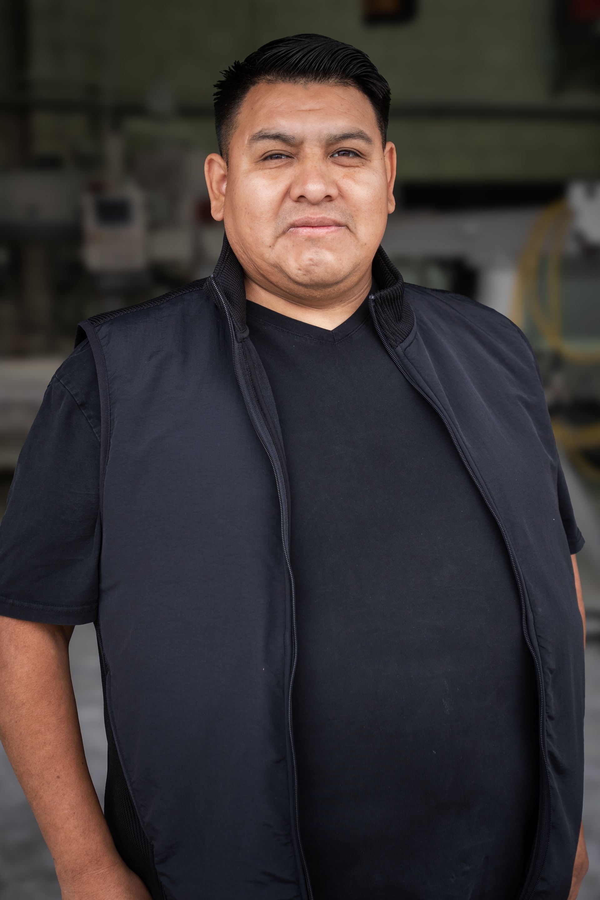 Person standing indoors wearing a black shirt and dark vest, facing the camera with a neutral expression