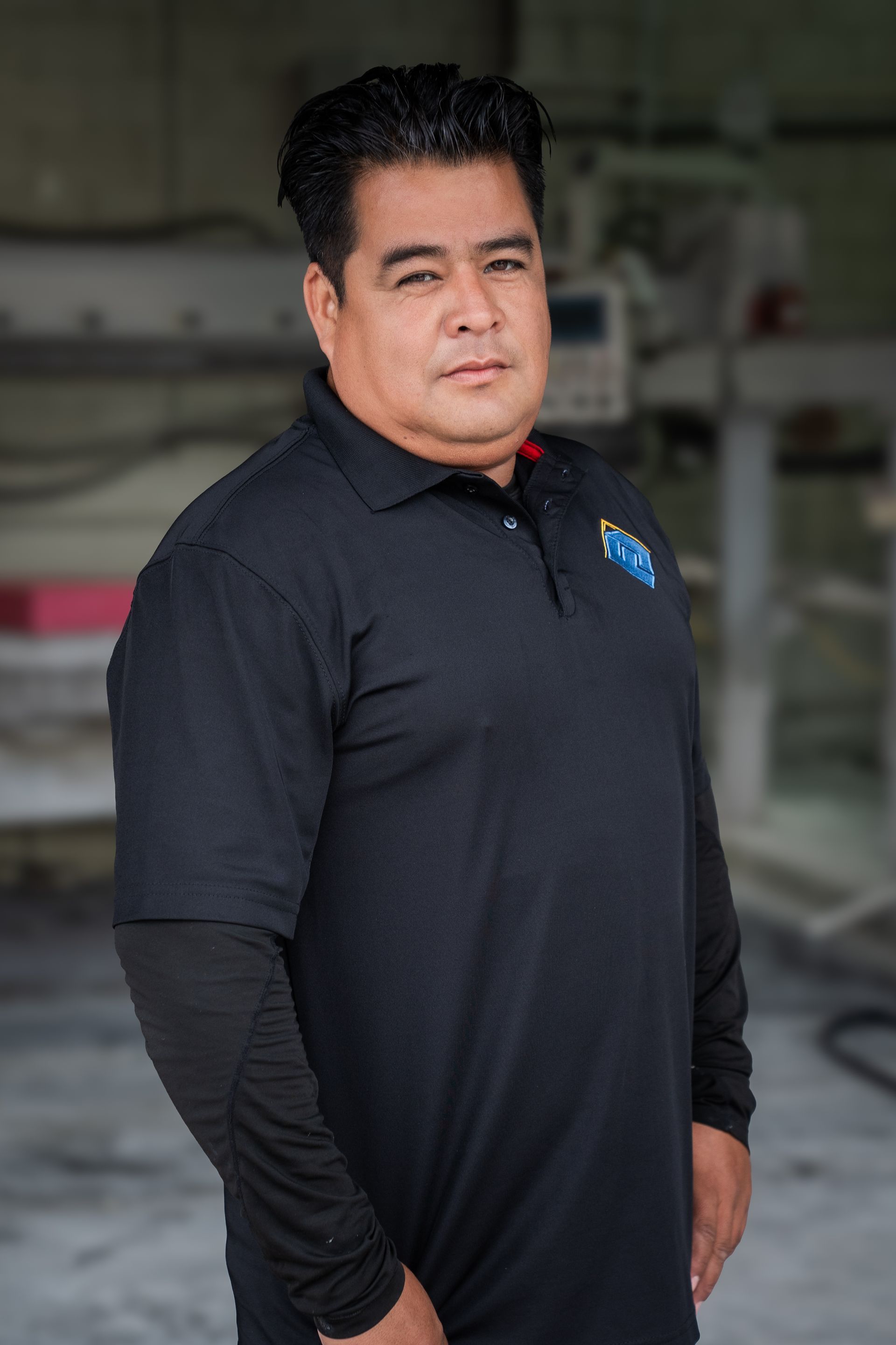 Man in a black polo shirt standing indoors with a neutral expression