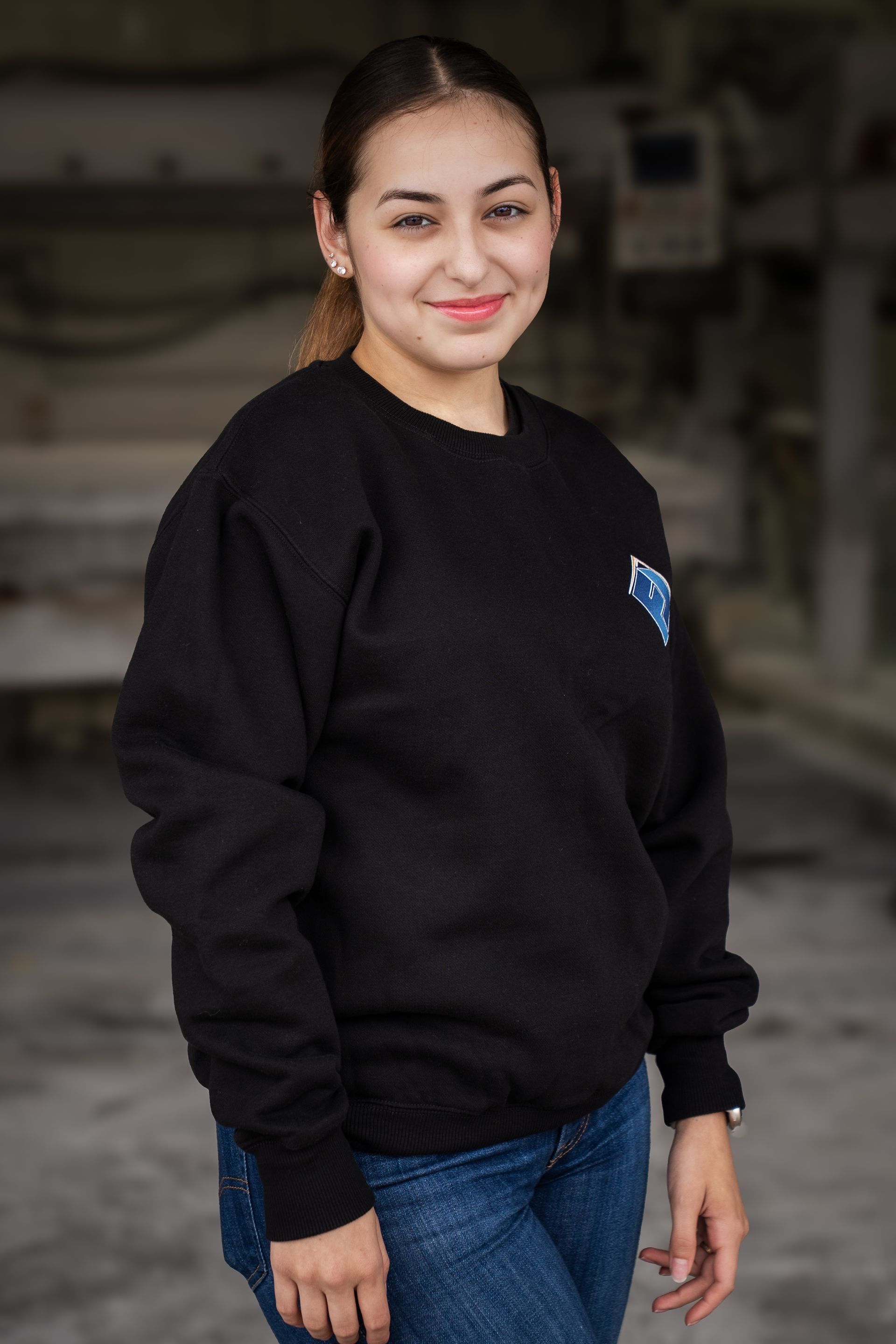 Woman standing indoors in a black sweatshirt and blue jeans, smiling at the camera.