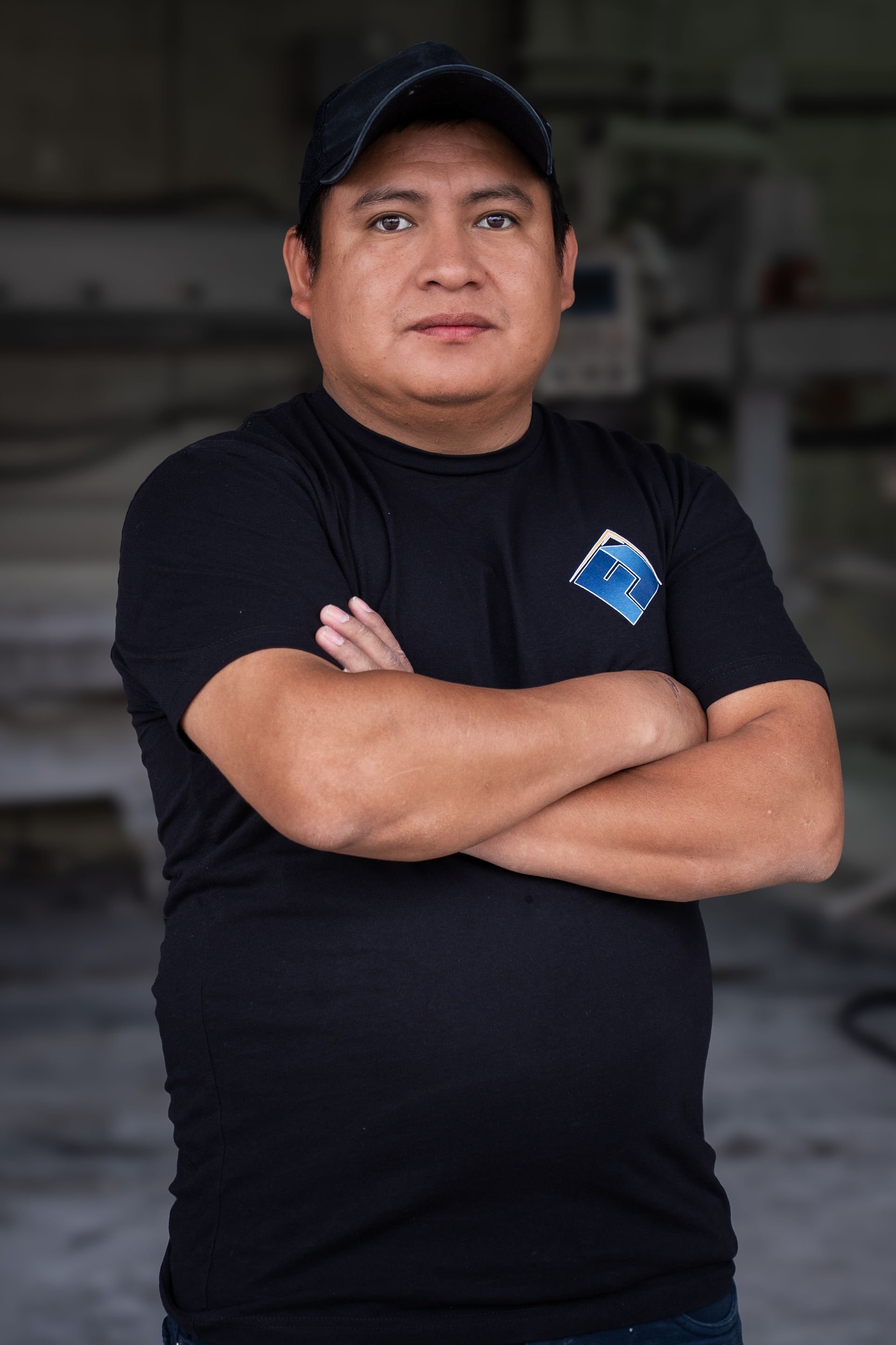 Man standing with arms crossed in a black T-shirt and cap, against a blurred indoor background