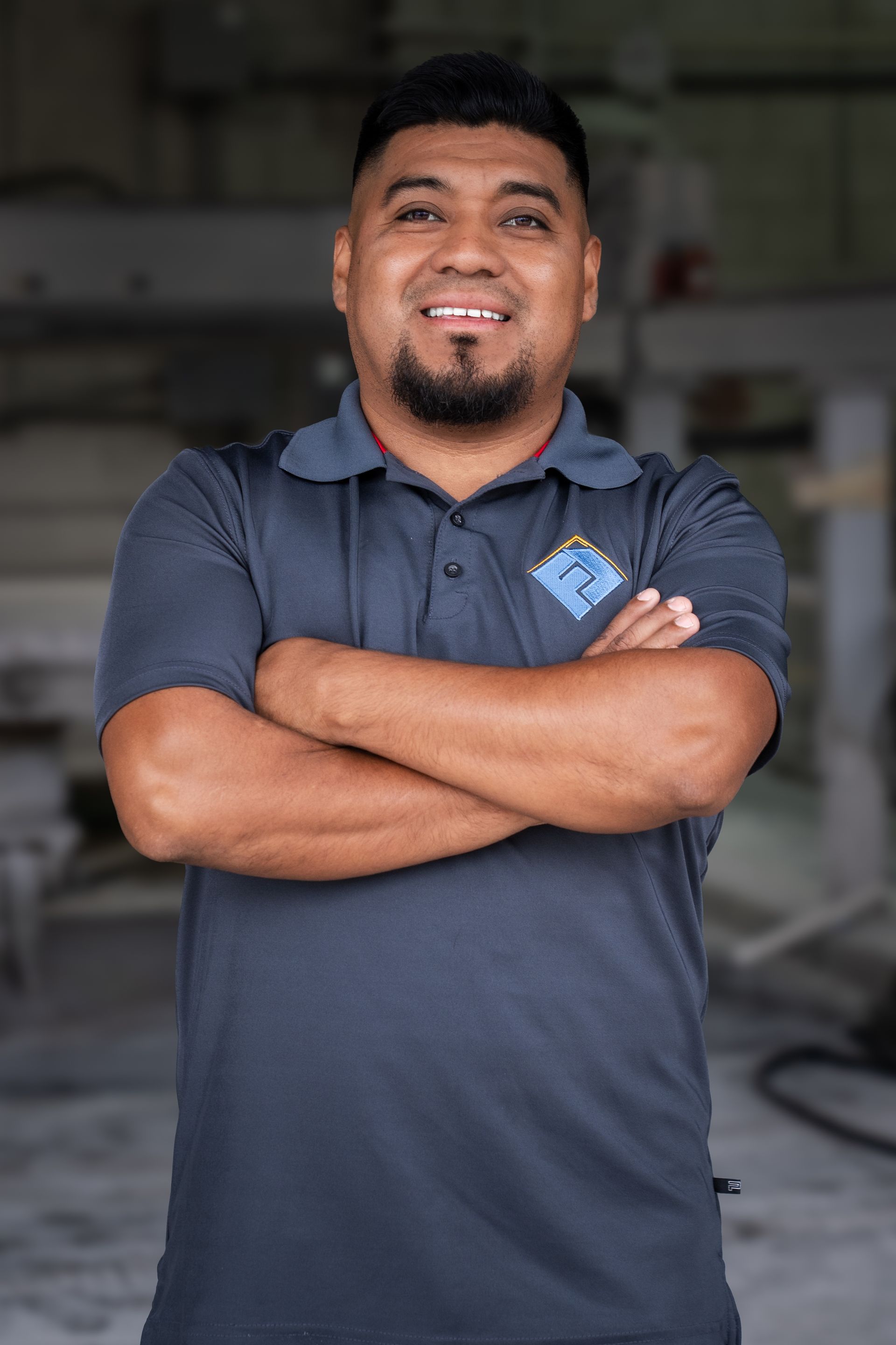 Smiling man in a navy polo shirt with arms crossed, standing indoors against a blurred background