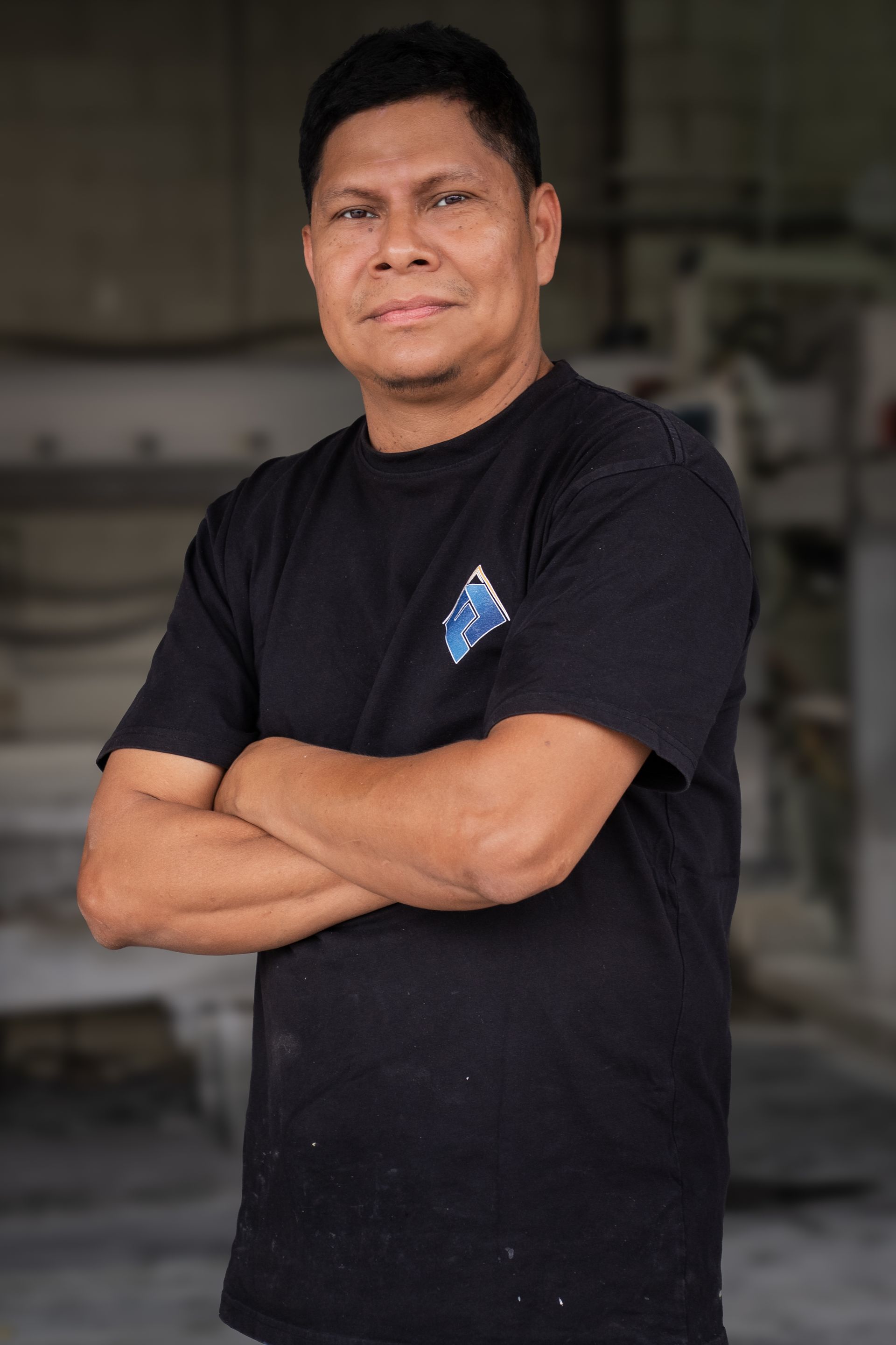 Man standing with arms crossed, wearing a black T-shirt in a workshop setting.