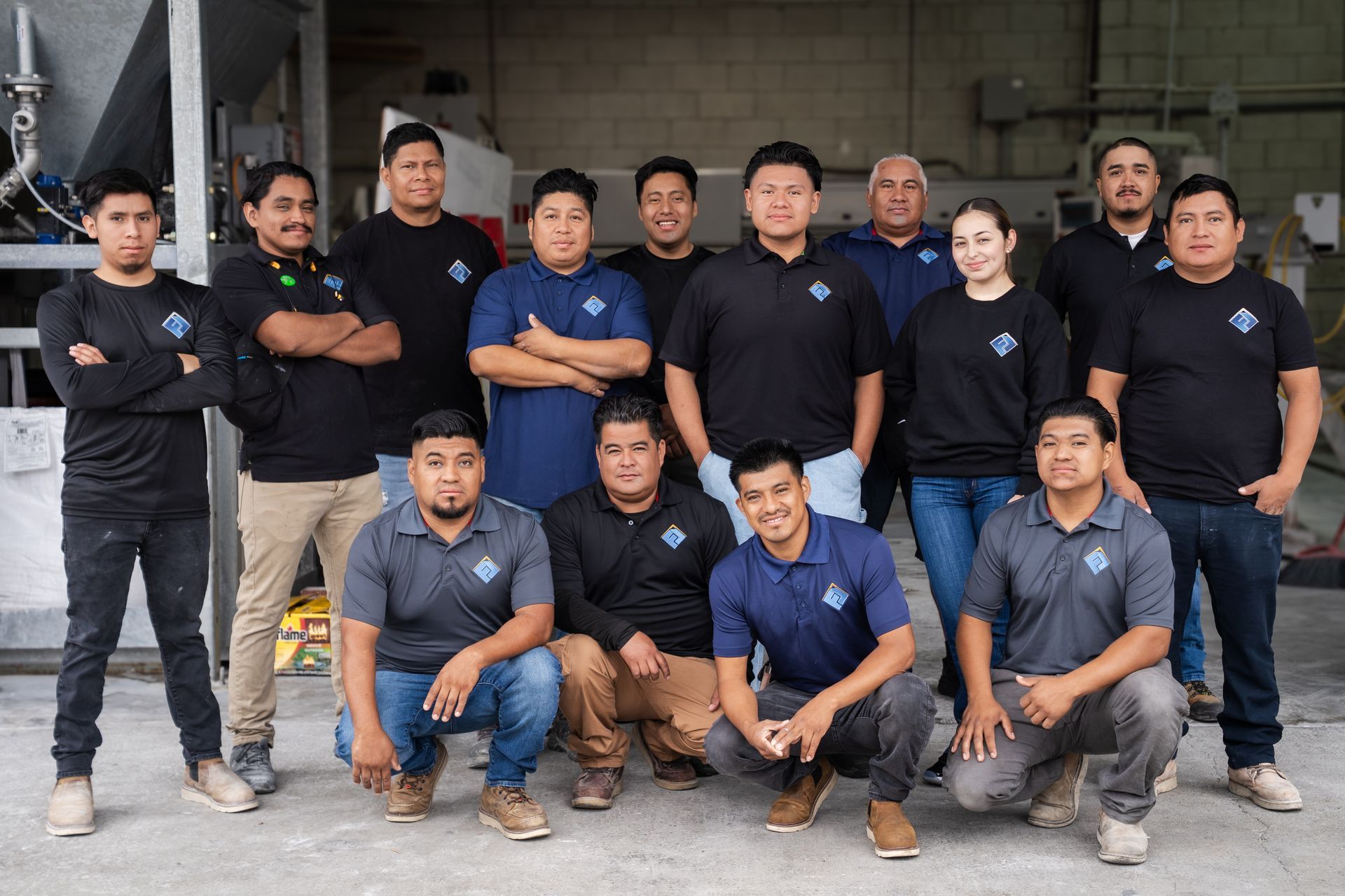 Group of men posing together indoors, some standing and some crouching, in matching dark shirts and work pants