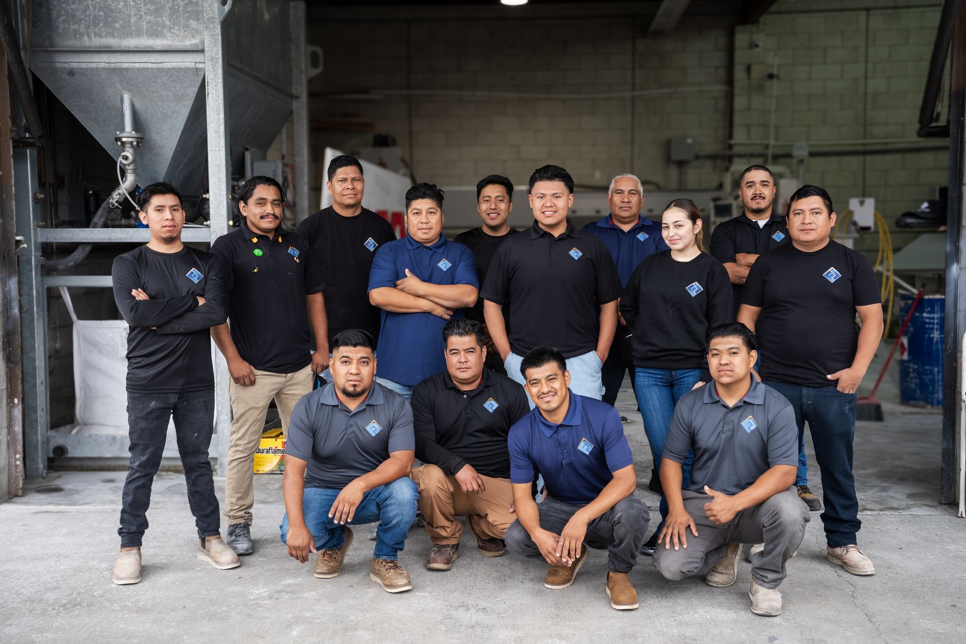 Group of people in dark shirts posing in front of an industrial building, some standing and some crouching.