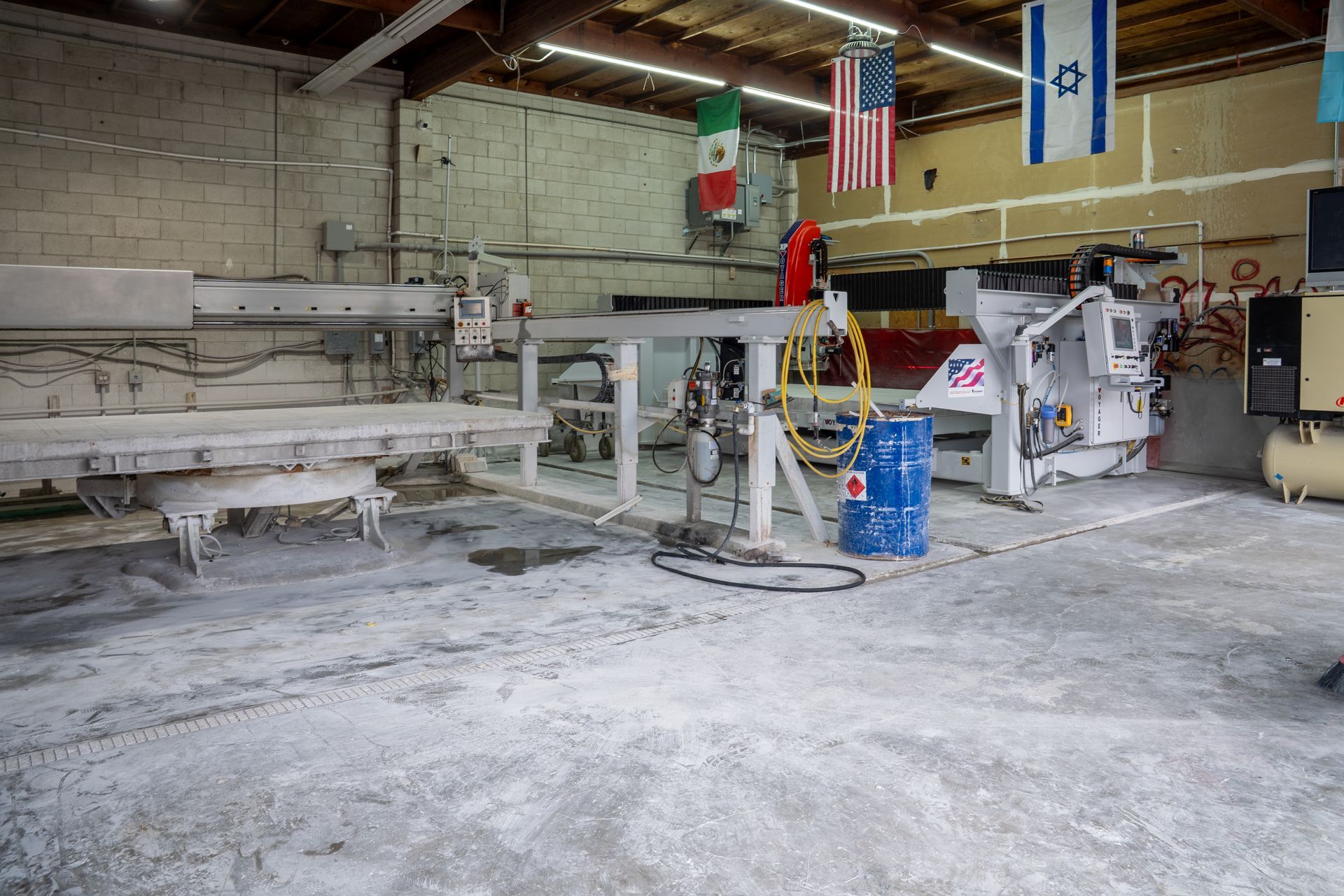 Industrial workshop with a machine, blue barrel, and hanging U.S. and Israeli flags, dusty floor.