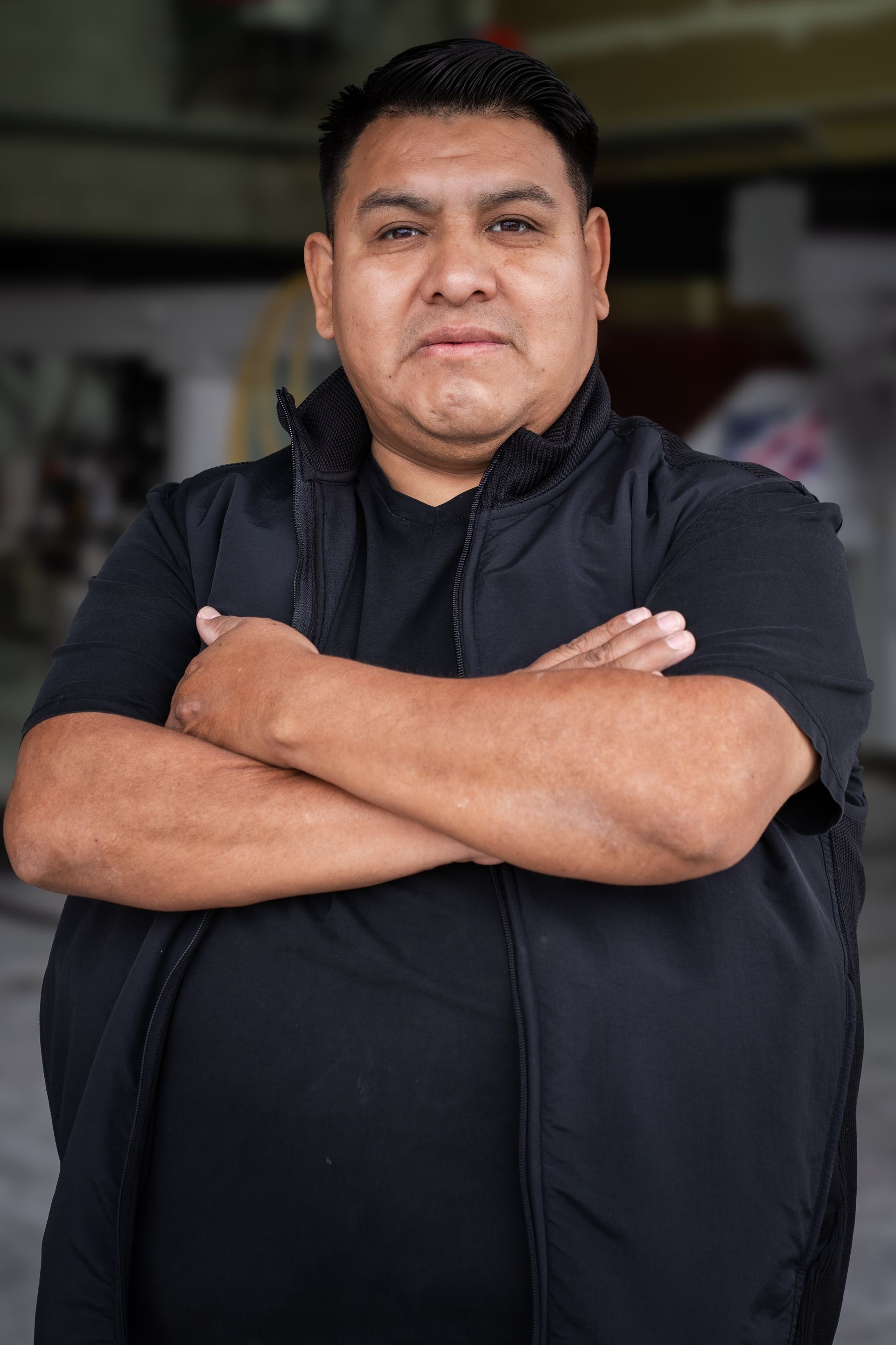 Person in a black jacket with arms crossed, standing indoors against a blurred background.