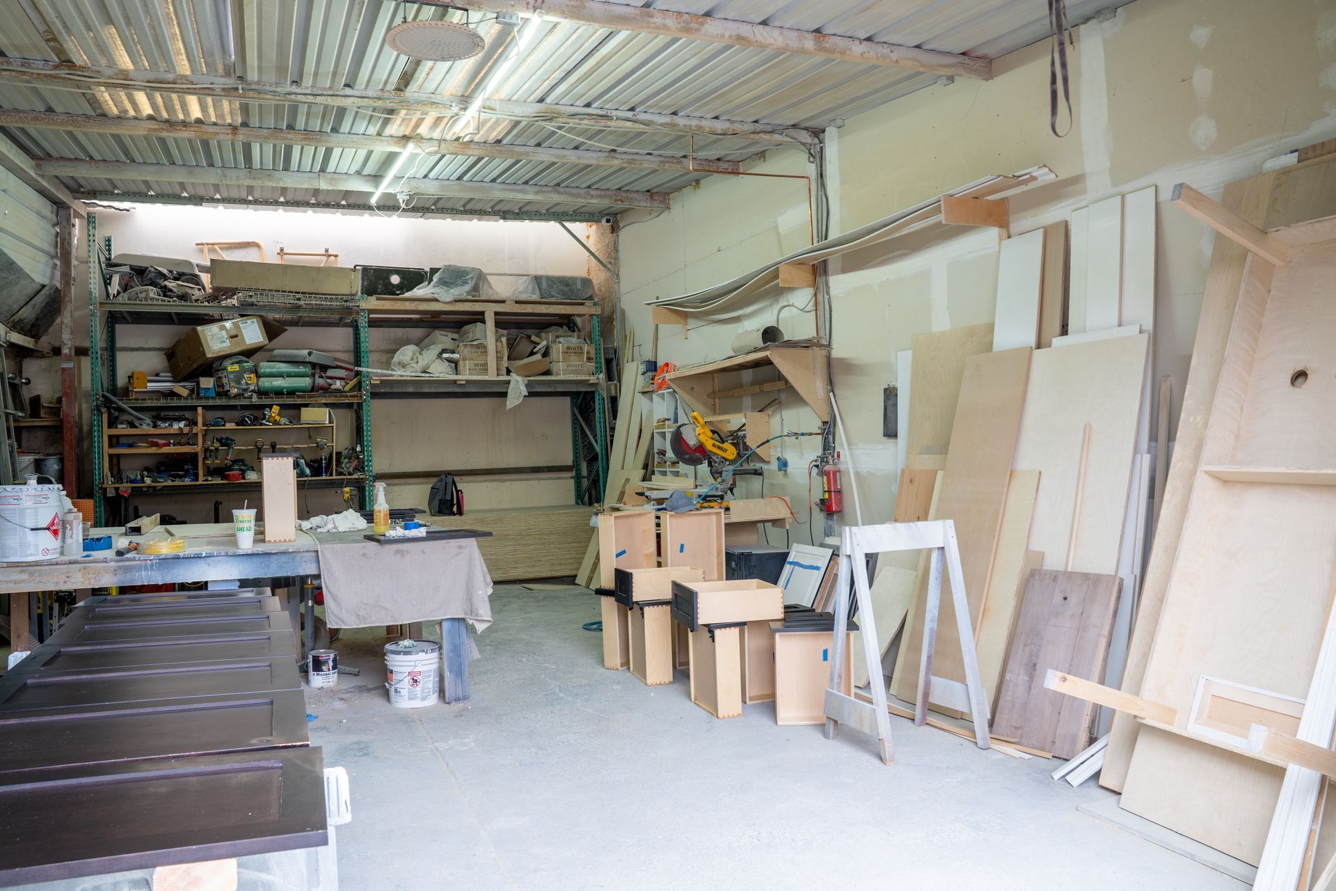Cluttered workshop interior with shelves, tools, cardboard boxes, and framed panels leaning against the wall