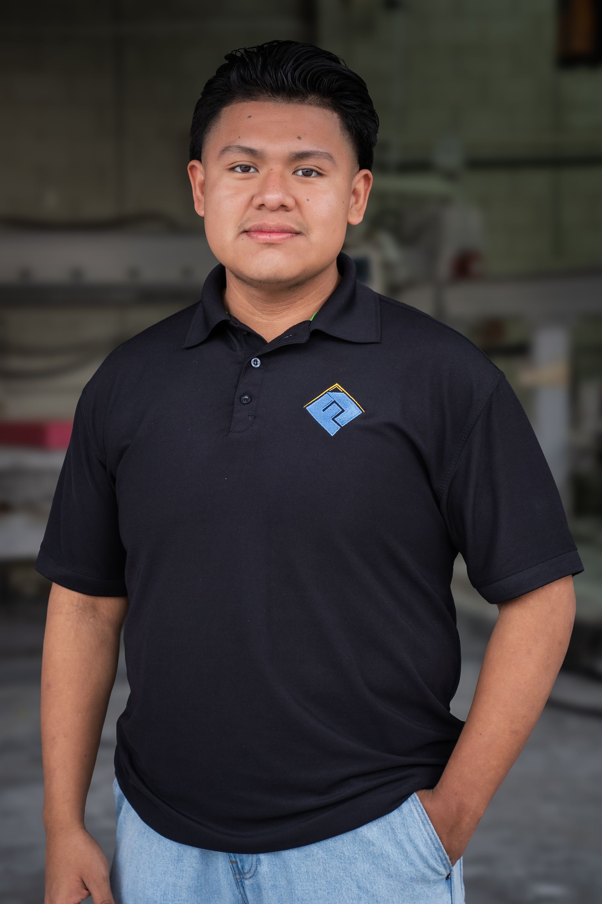 Person standing indoors wearing a black polo shirt and blue jeans, with one hand in a pocket.