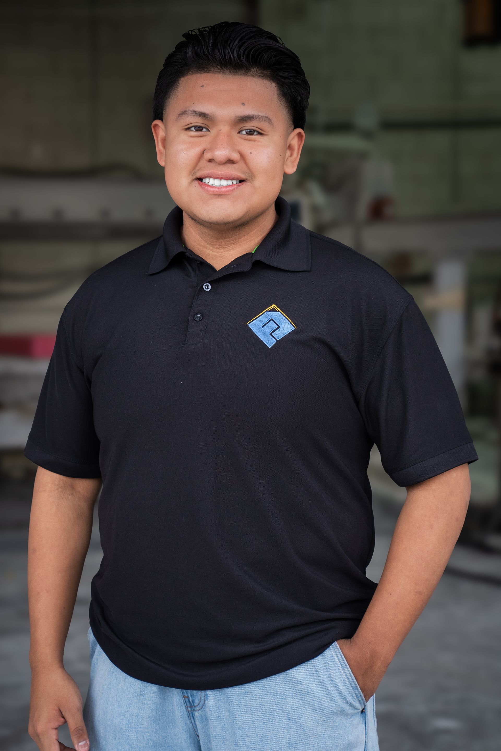 Smiling person in a black polo shirt and light jeans standing indoors in front of a blurred background