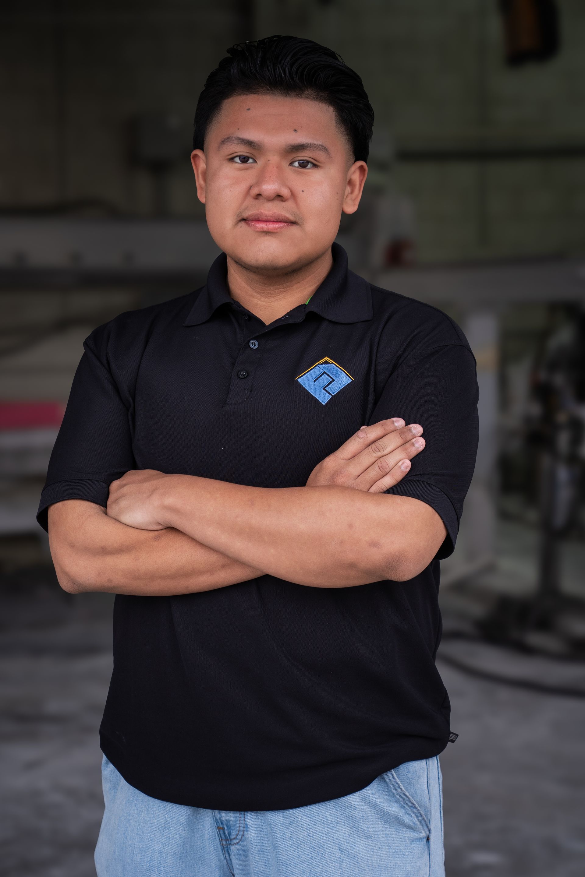 A man in a black polo shirt stands with arms crossed in a dim indoor setting.
