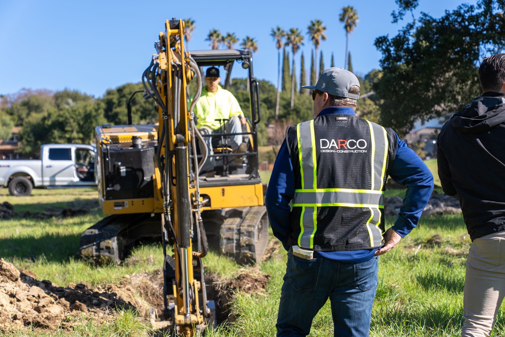 Three men on a construction site. One operates an excavator, while two others watch, wearing safety vests.