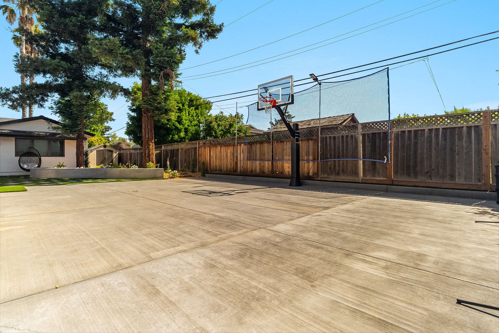 Empty concrete backyard with a basketball hoop, wooden fence, and tall trees under a clear blue sky