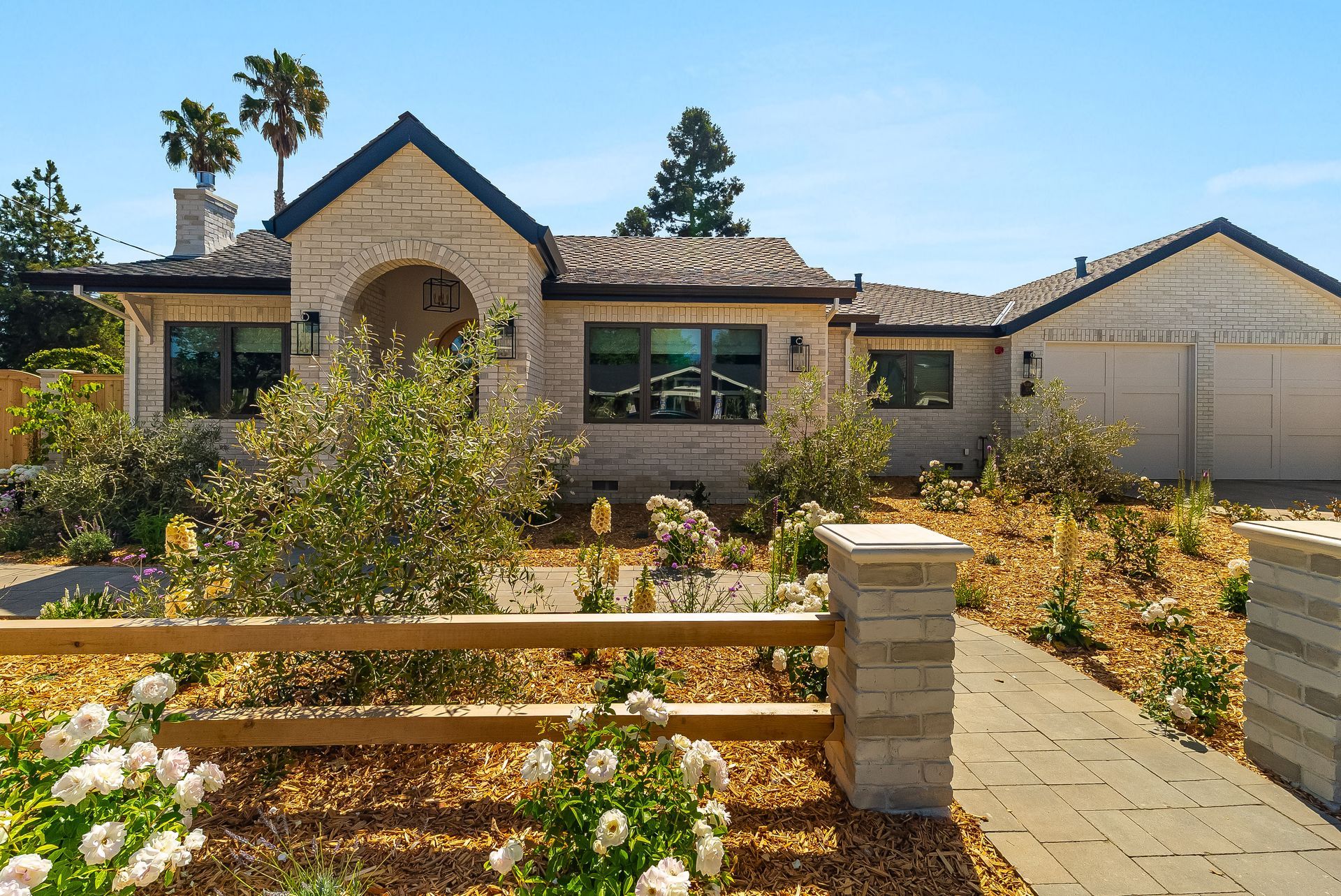 Front yard of a beige house with stone accents, white flowers, and a paved walkway on a sunny day