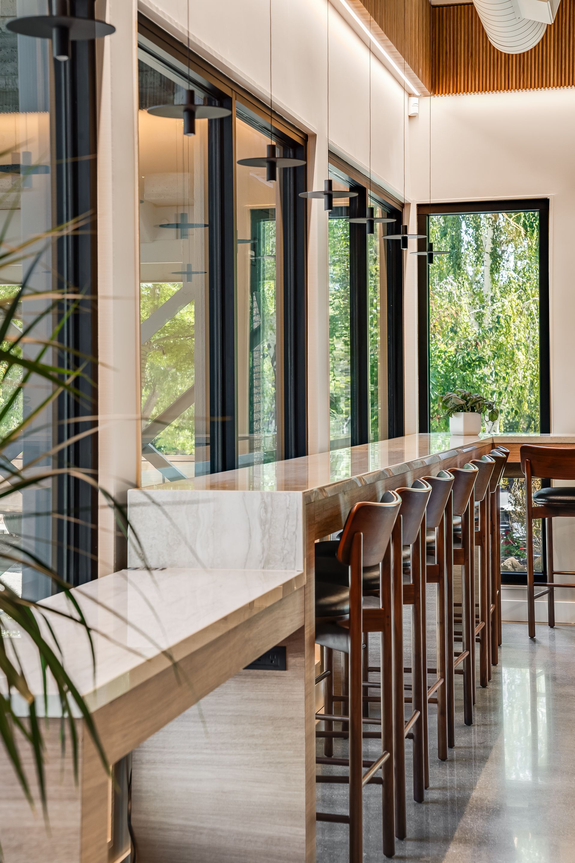 Modern café interior with tall wooden stools by a marble counter and large windows overlooking greenery