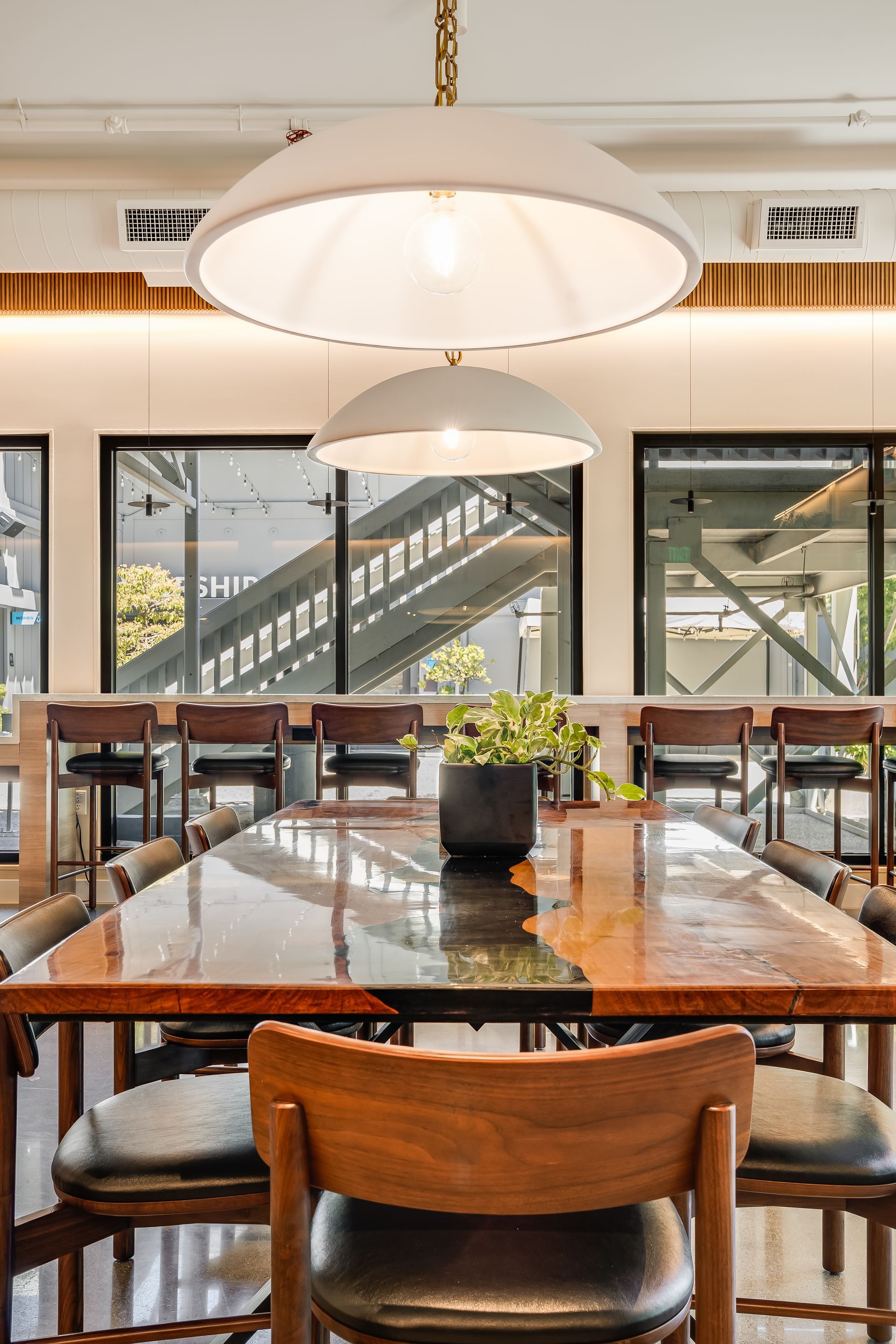 Bright cafe interior with wooden tables, large pendant lights, and windows overlooking an outdoor stairway
