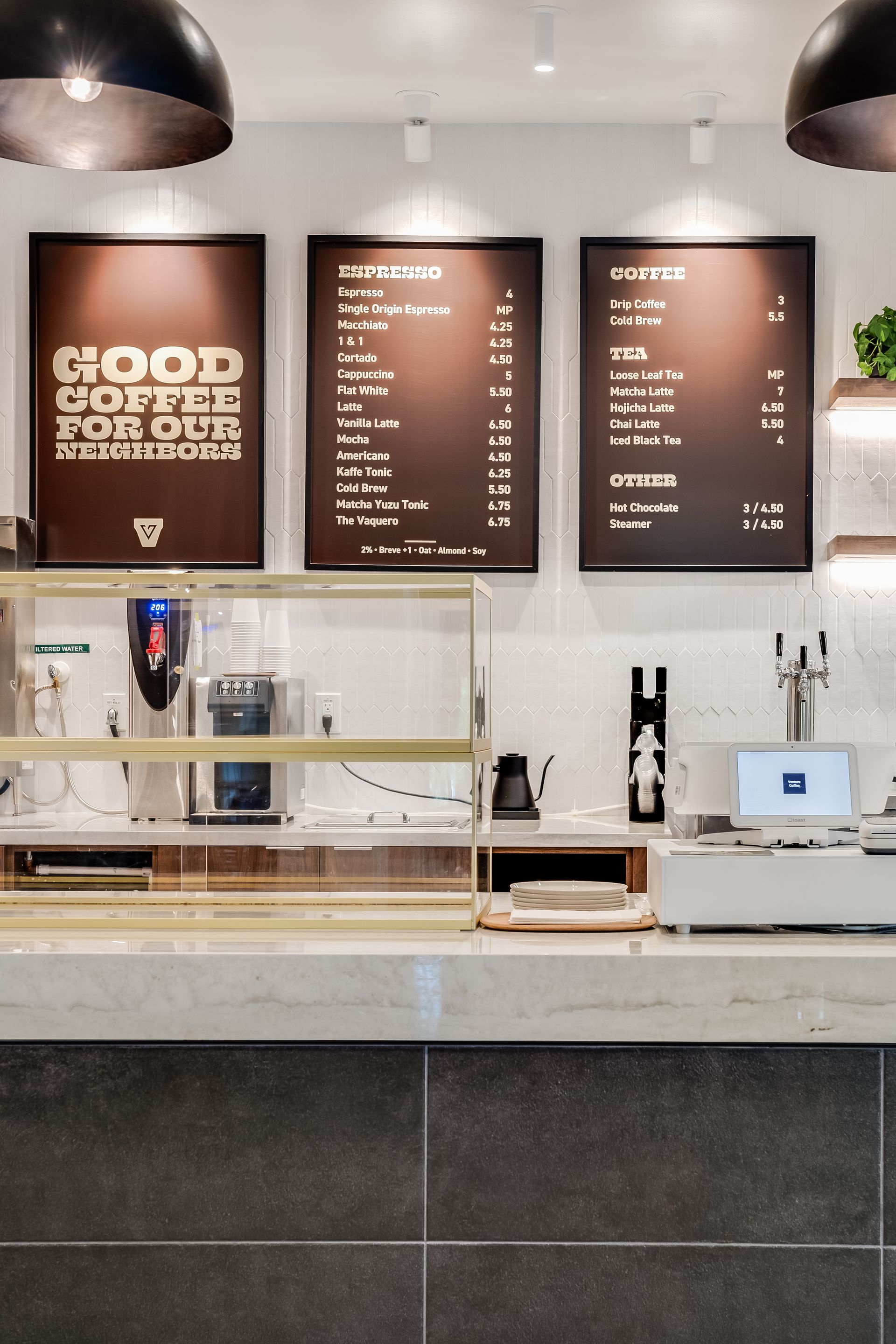 Modern café counter with menu boards, espresso machine, and glass display case behind a tiled front counter